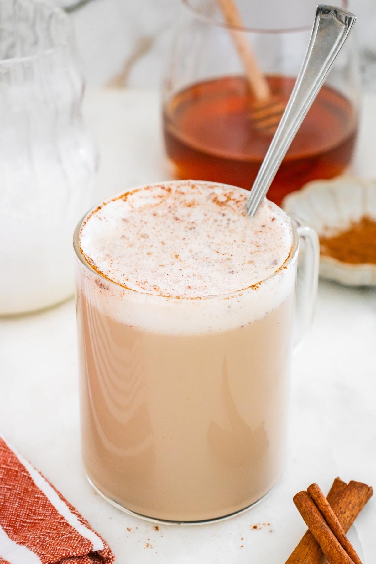 A glass mug of chai latte with frothy milk, topped with cinnamon, and a spoon inside.