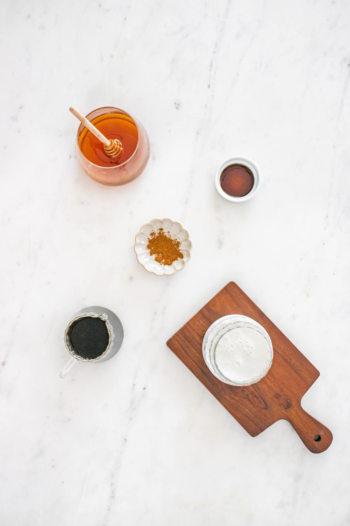 Ingredients on a marble surface: honey, cinnamon, coffee, vanilla, and a glass of milk on a wooden board.