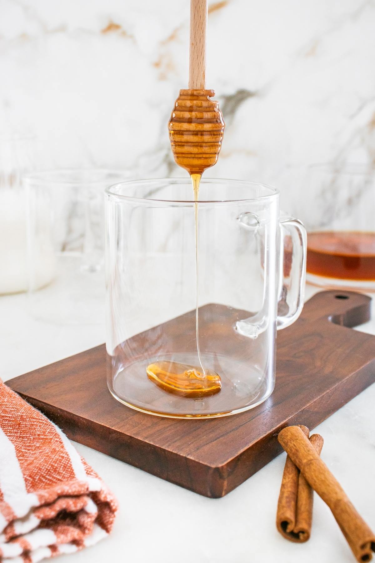 A honey dipper drizzling honey into an empty glass mug on a wooden board with cinnamon sticks nearby.