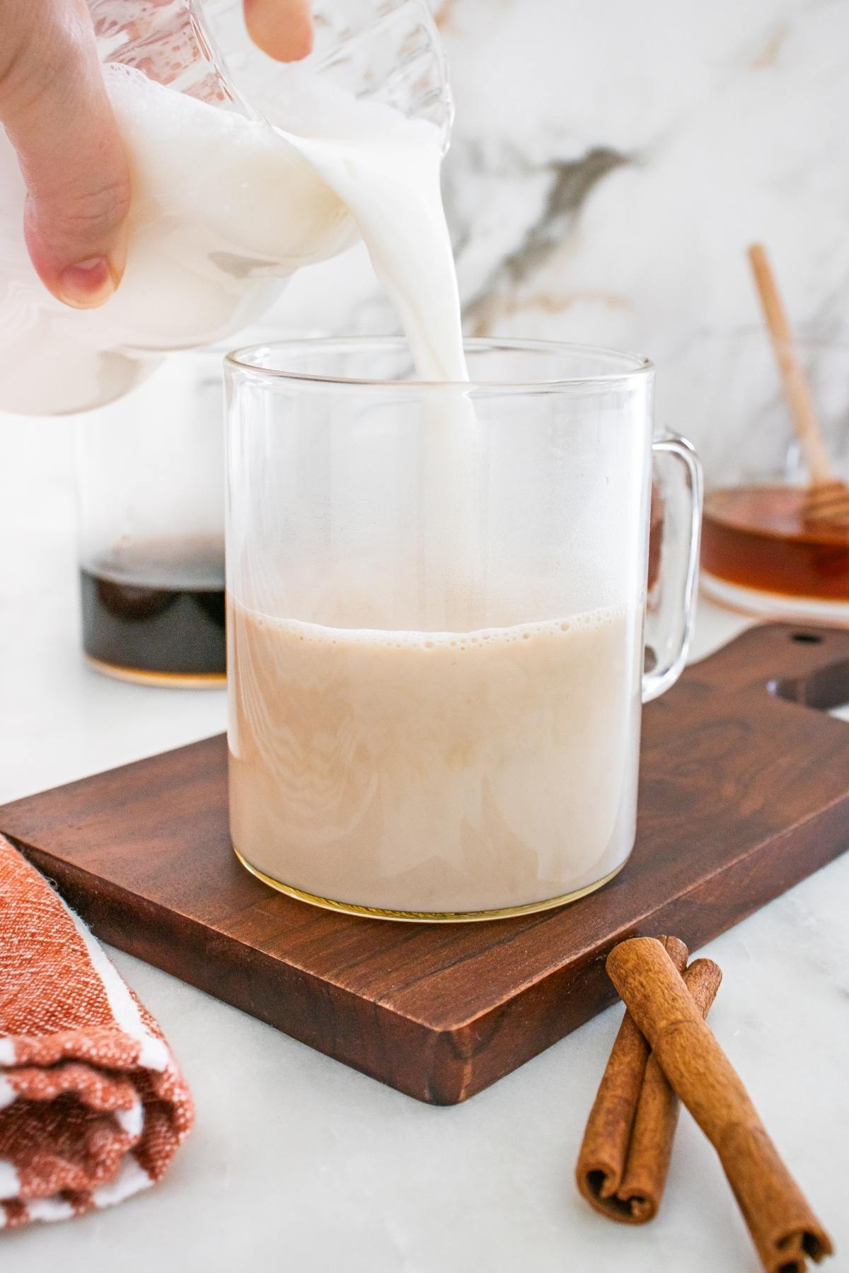 Milk is being poured into a glass mug on a wooden board, with cinnamon sticks and honey in the background.