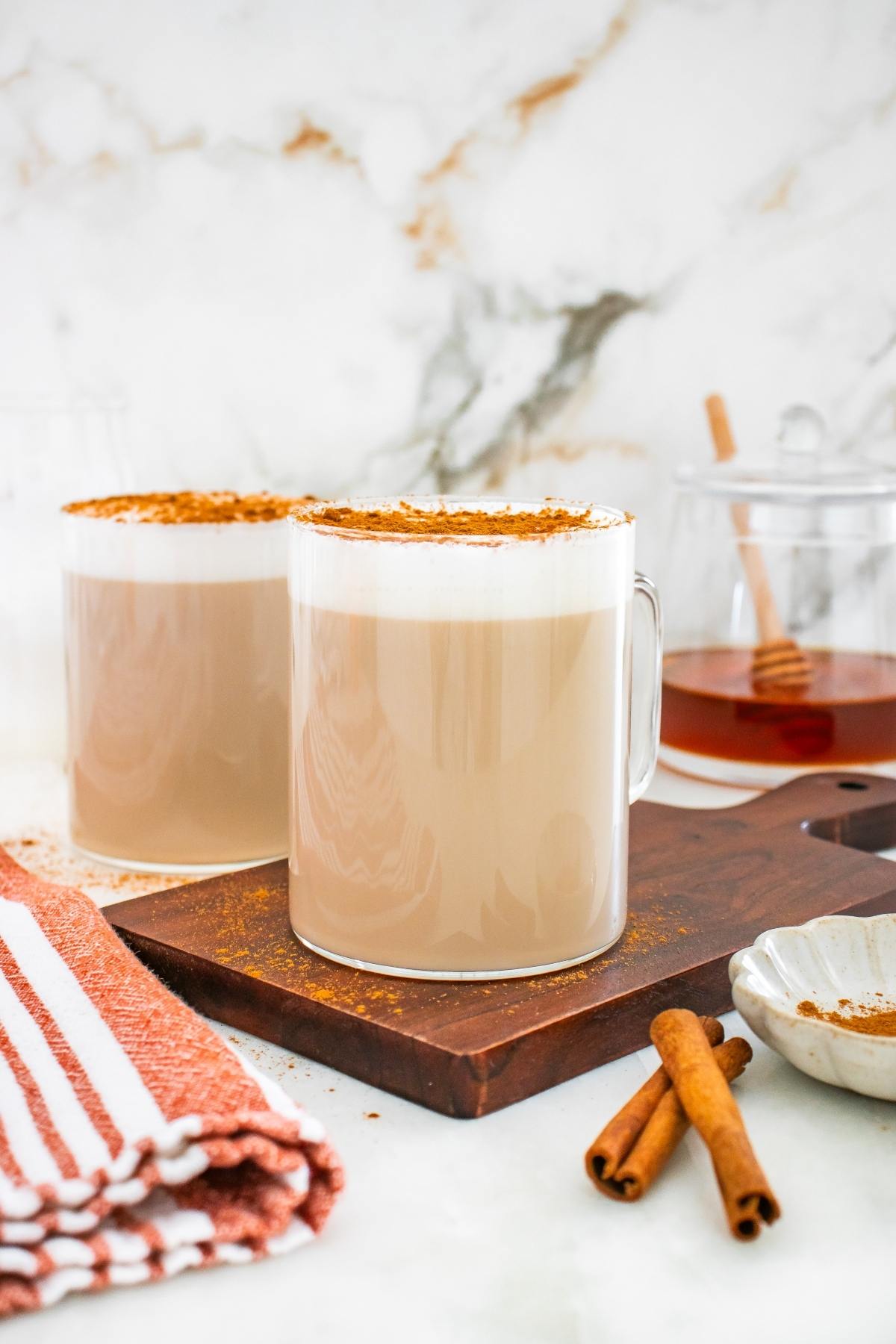 Two mugs of frothy chai latte topped with cinnamon sit on a wooden board, with honey and cinnamon sticks nearby.