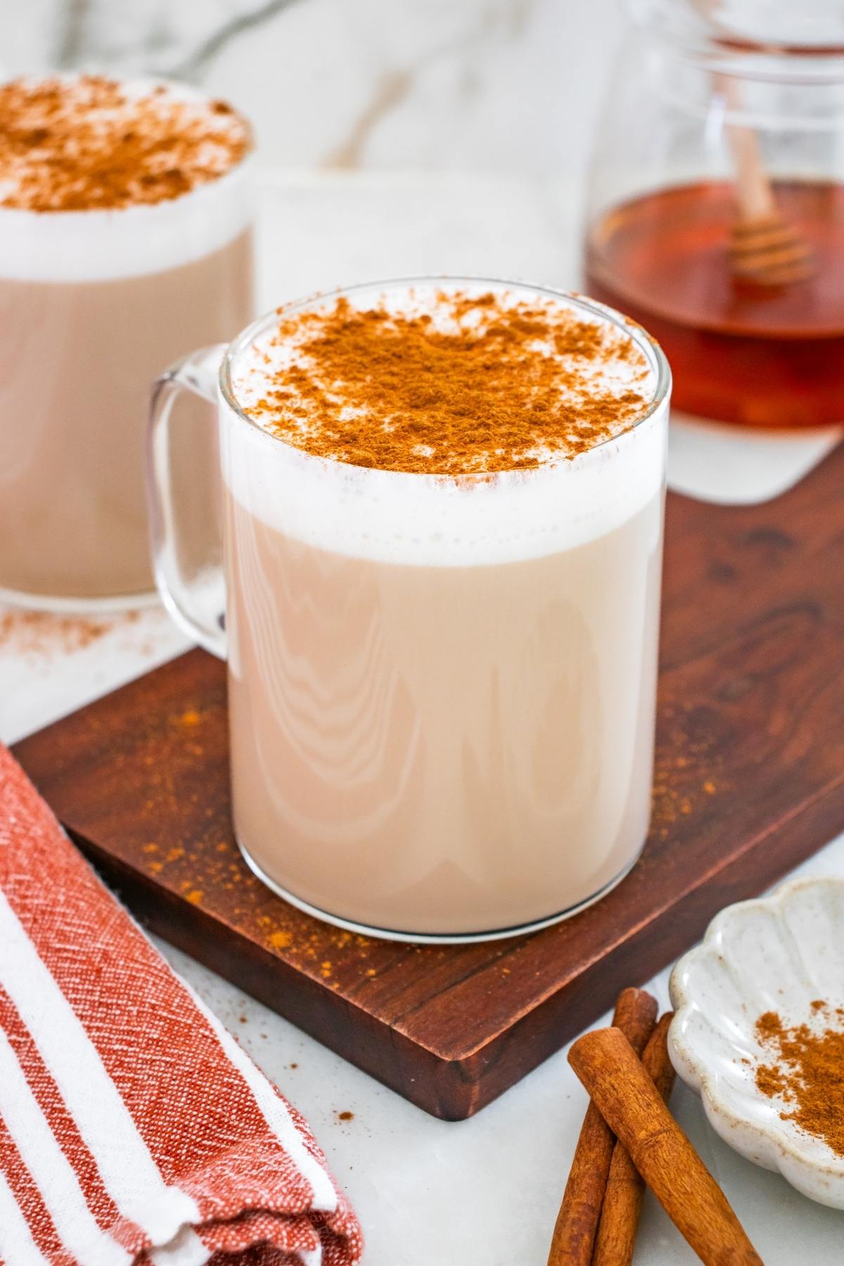 A glass mug of chai latte with frothy milk and cinnamon on top, next to cinnamon sticks and honey.