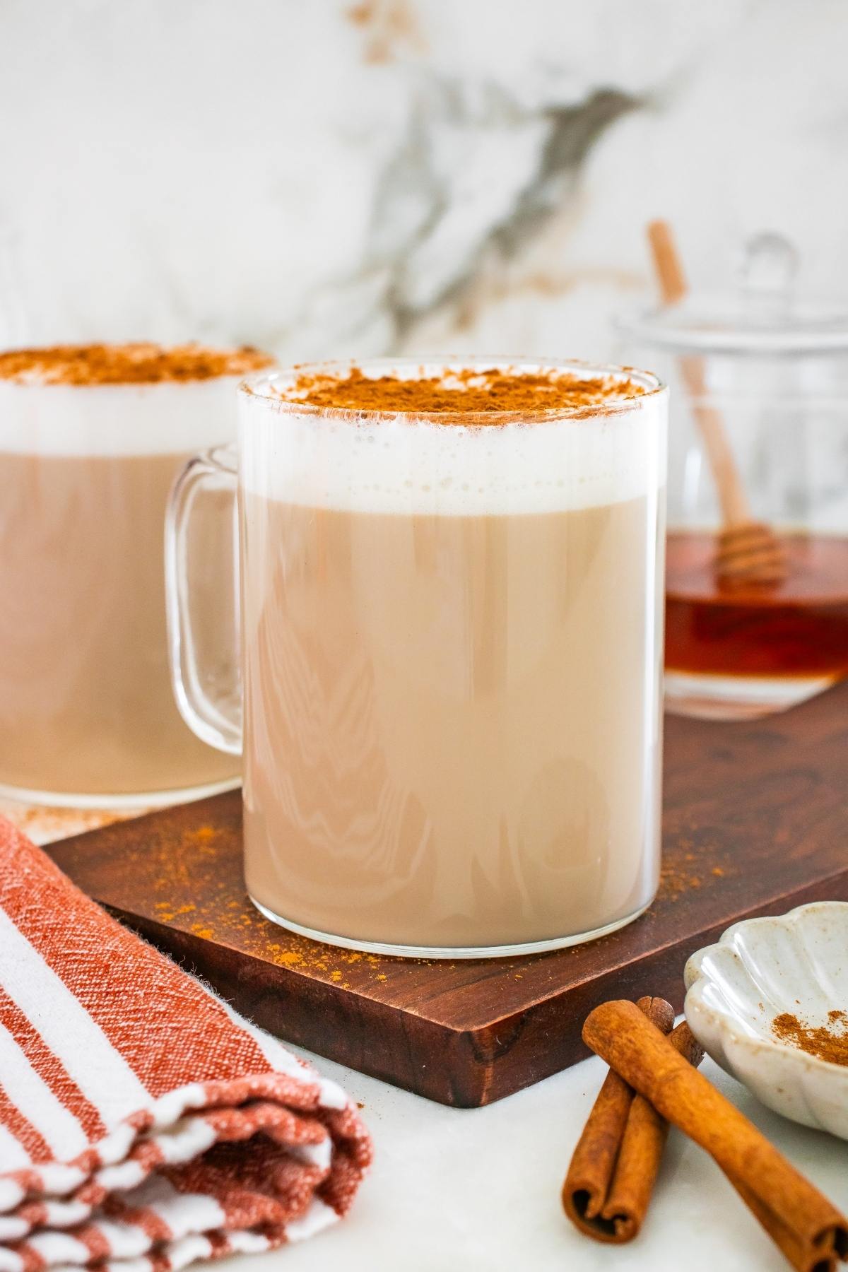 A glass mug of frothy chai latte topped with cinnamon, beside a napkin and cinnamon sticks on a wooden board.