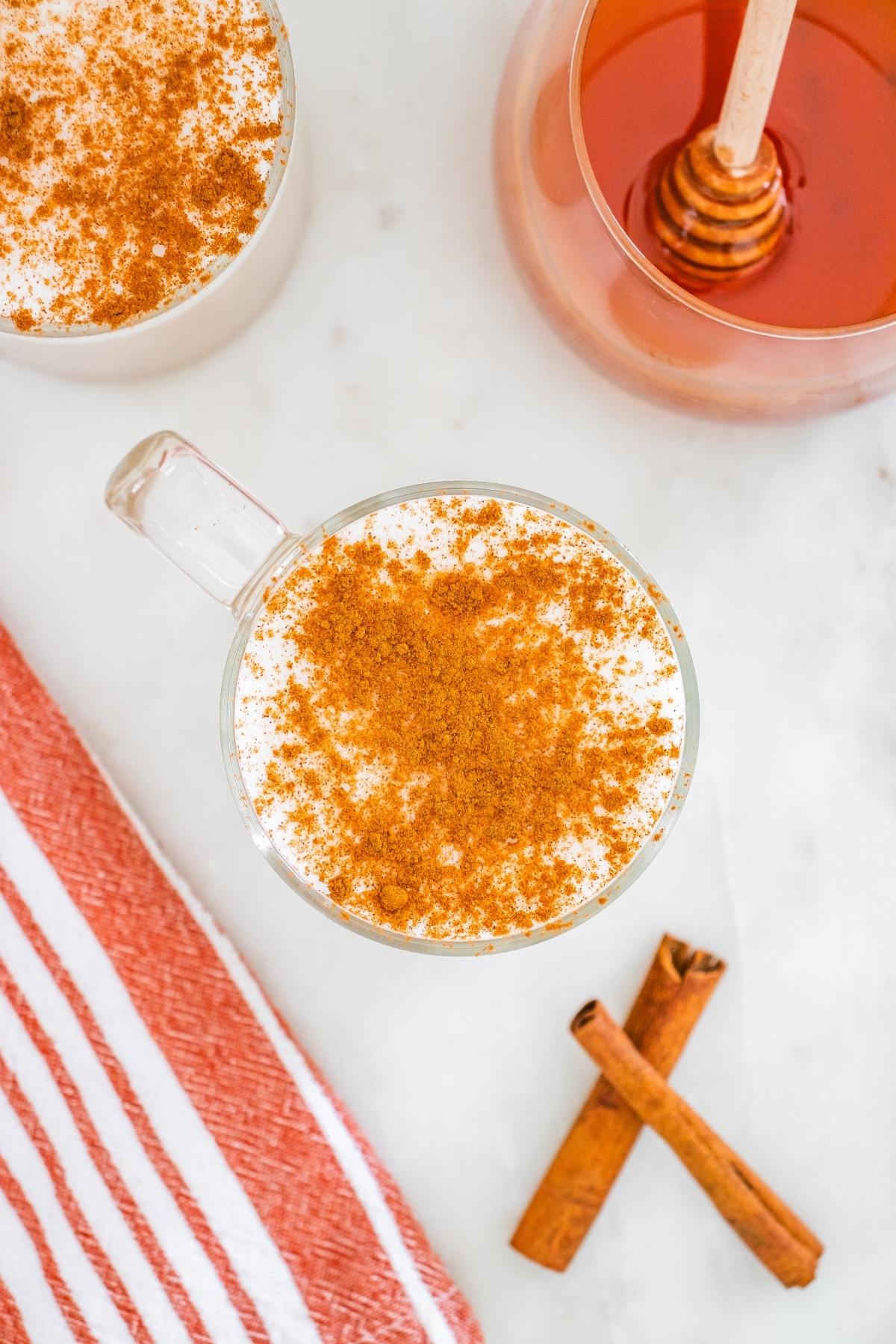 A mug of frothy drink topped with cinnamon, next to cinnamon sticks, honey, and a red striped towel.