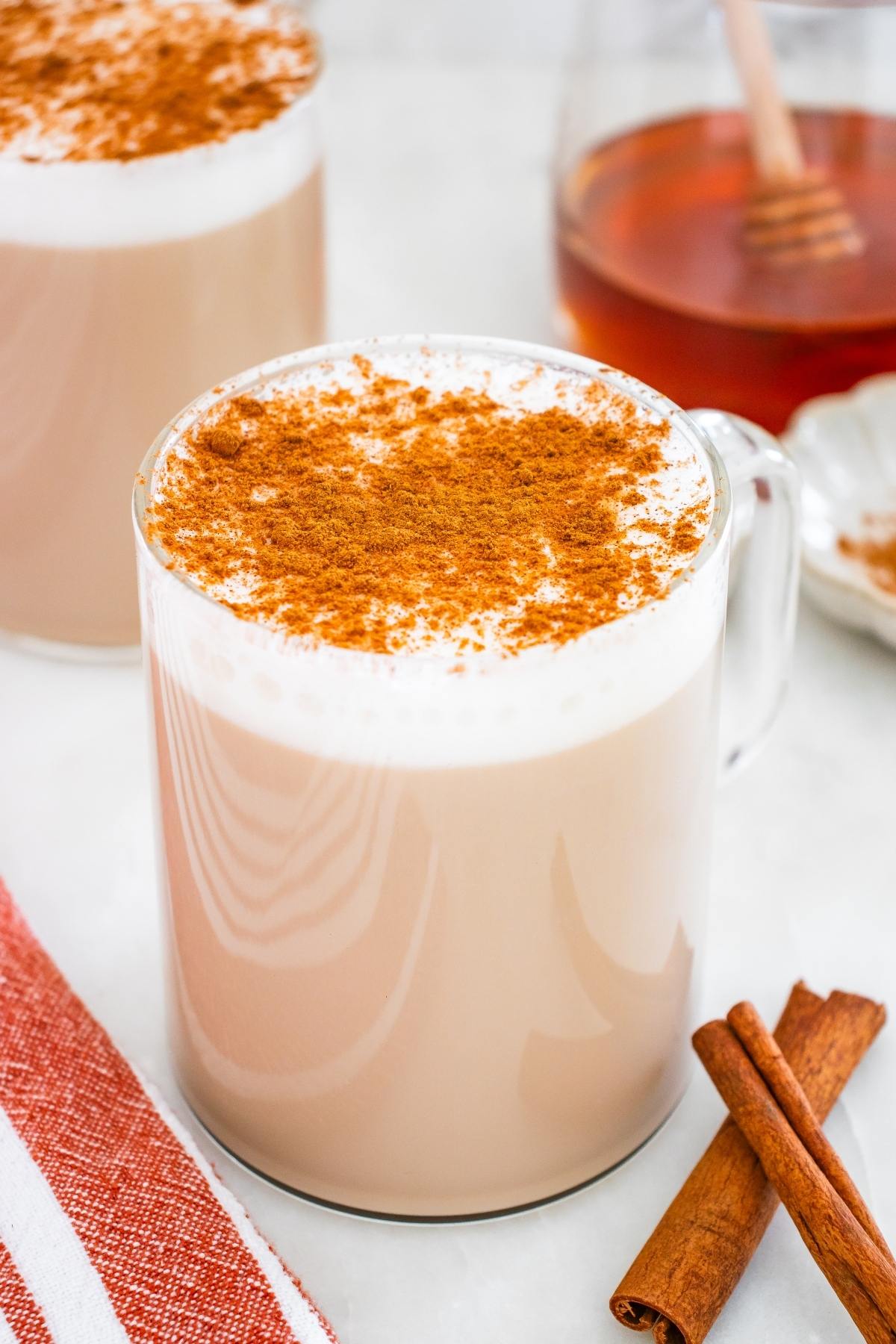 A mug of chai latte topped with cinnamon, beside cinnamon sticks and a jar of honey.