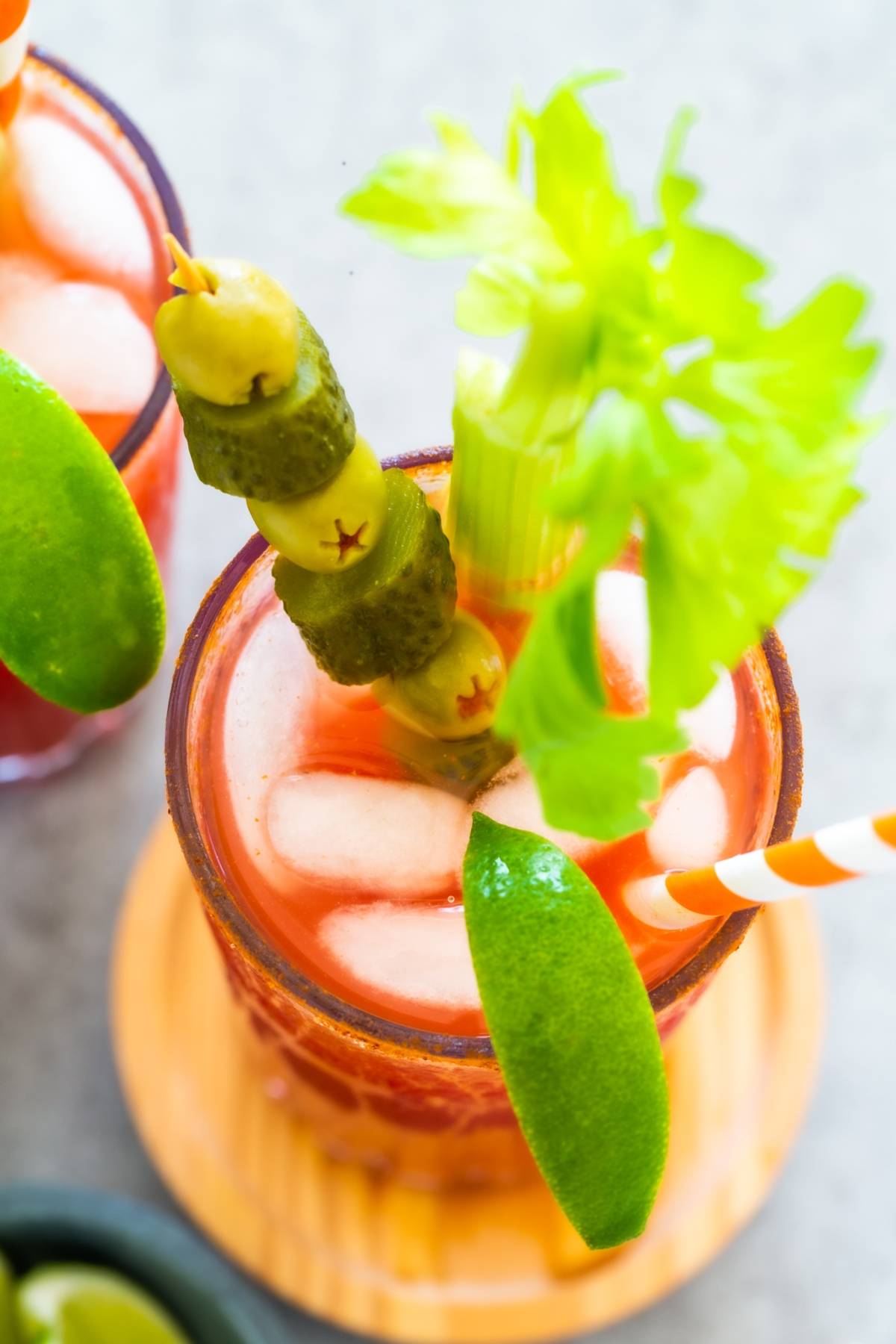 Bloody Mary cocktail with ice, celery stalk, pickles, lime wedge, and striped straw—this Canadian Caesar Cocktail is viewed from above.