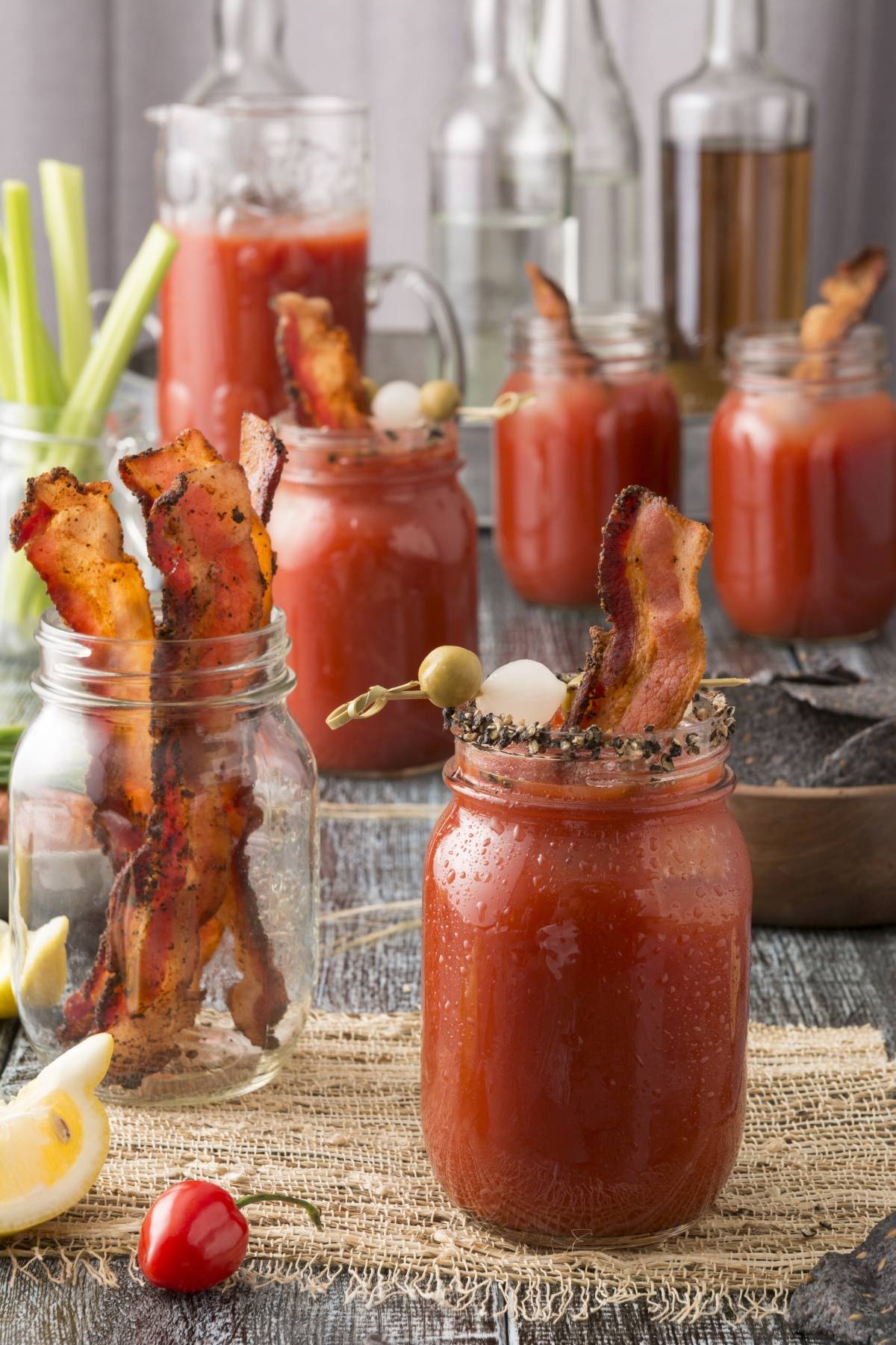 Mason jars of Canadian Caesar Cocktail, garnished with bacon, olives, and onions on a rustic table—a perfect cocktail for Canada Day.