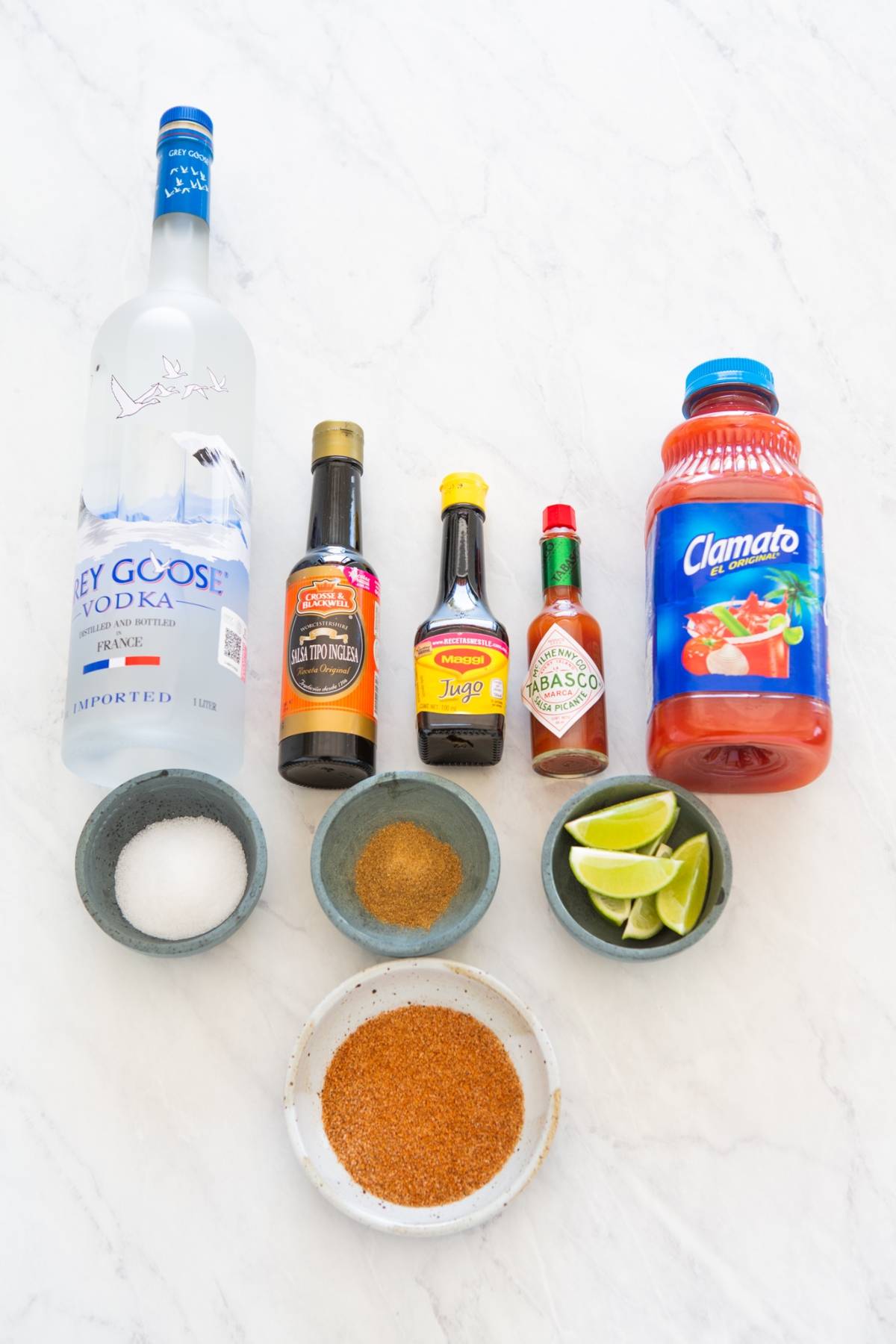 Bottles of vodka, Clamato, sauces, lime wedges, sugar, and spices arranged on a white surface—perfect caesar cocktail ingredients for a classic bloody mary with Clamato juice or a festive cocktail for Canada Day.