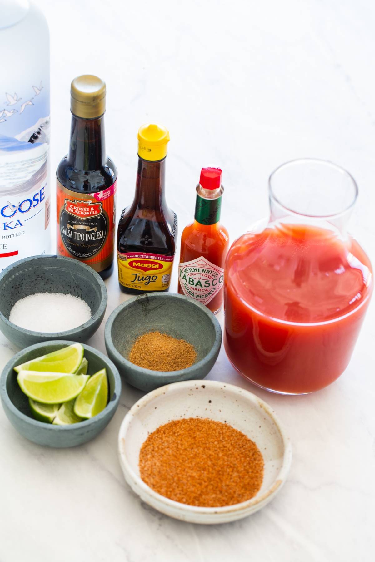 Various Caesar cocktail ingredients, including vodka, sauces, lime wedges, spices, and tomato juice on a white surface—everything you need to craft Canada's National Drink, the Canadian Caesar Cocktail.