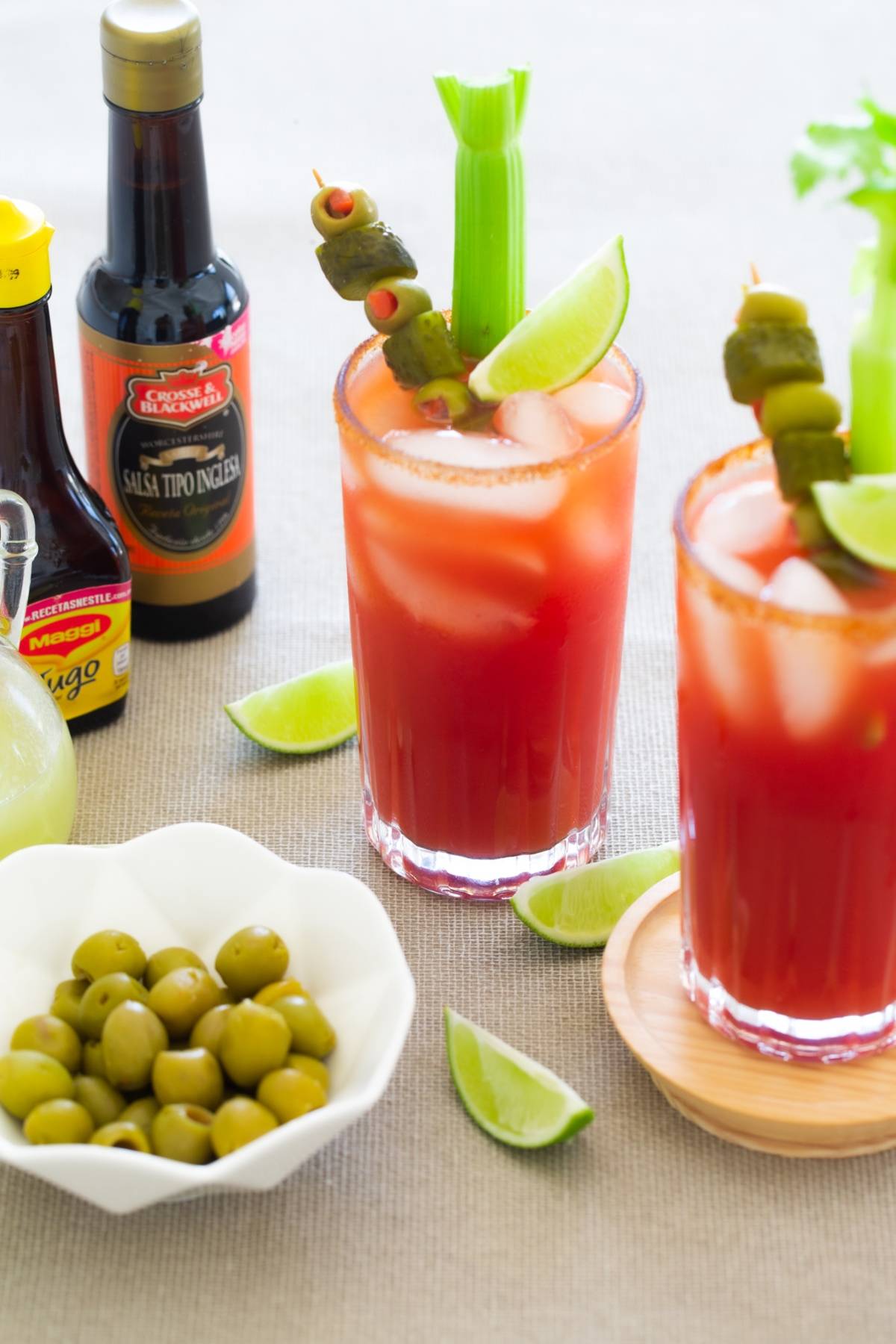 Two Bloody Mary cocktails with celery, lime, and olives, surrounded by sauces, lime wedges, and a bowl of olives—perfect as Canadian Caesar cocktail ingredients or as a festive cocktail for Canada Day.
