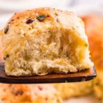 A wooden spatula holds a fluffy, golden Cheddar Chive Dinner Roll above a baking dish filled with similar herb-topped rolls.