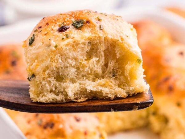A wooden spatula holds a fluffy, golden Cheddar Chive Dinner Roll above a baking dish filled with similar herb-topped rolls.