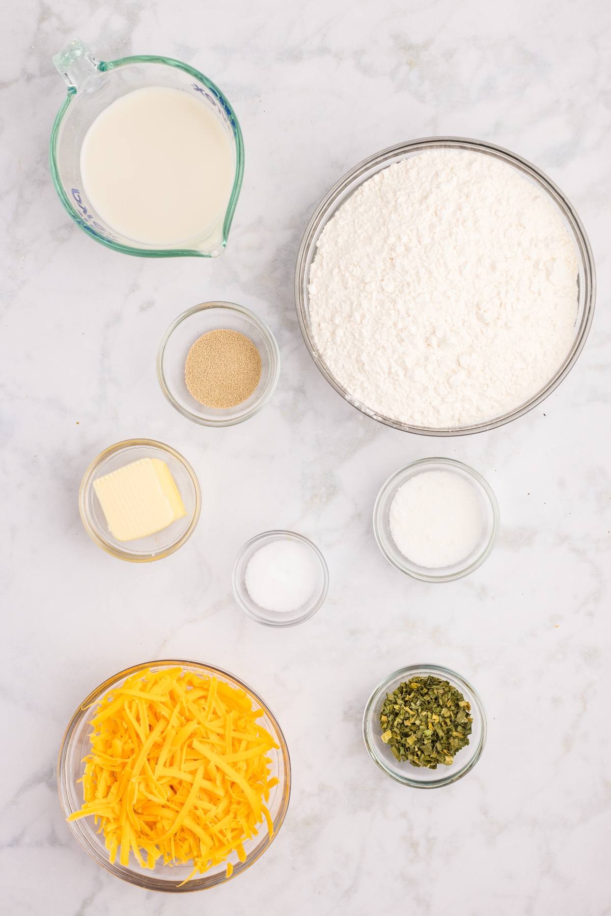 Bowl of flour, shredded cheese, butter, milk, yeast, sugar, salt, and herbs on a white marble surface.