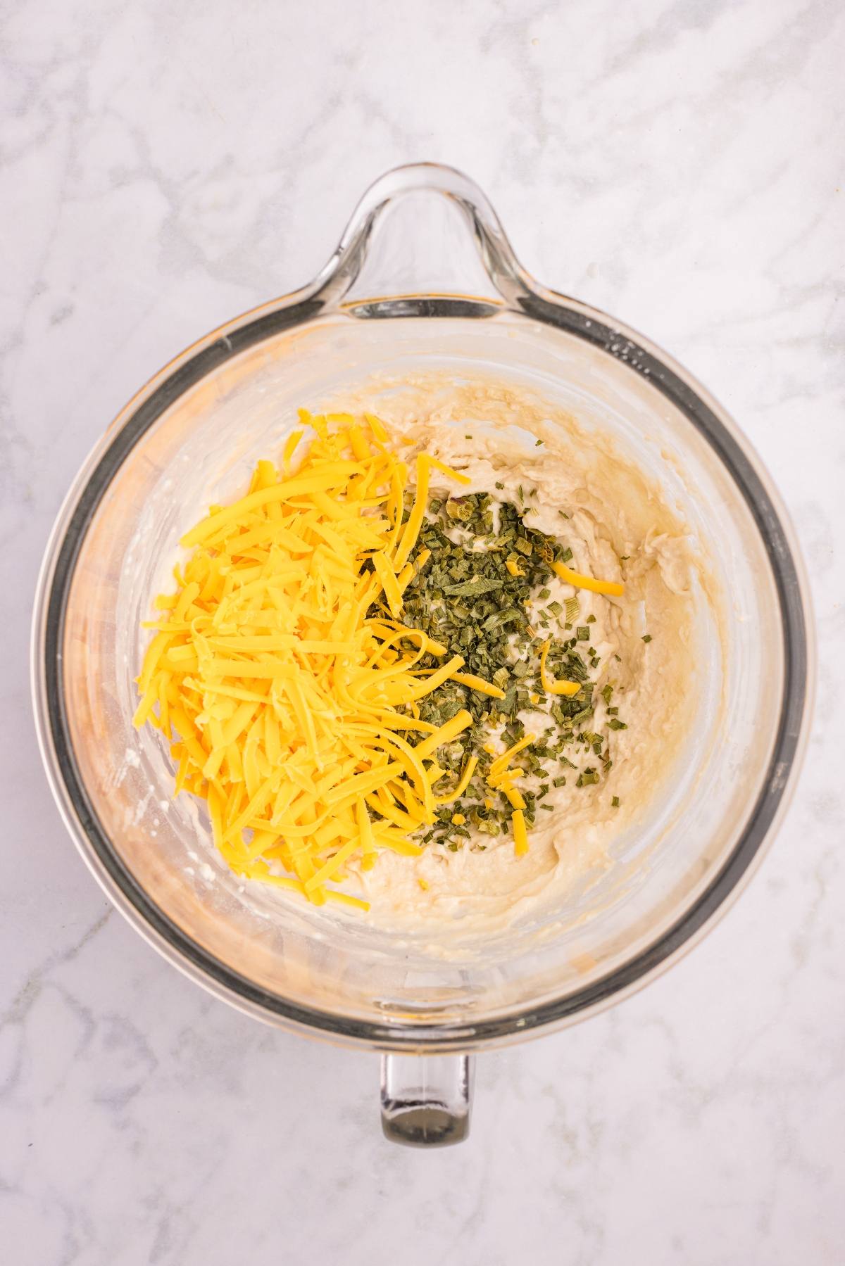 Glass bowl with shredded cheddar cheese and chopped herbs on top of a thick batter, on a marble surface.