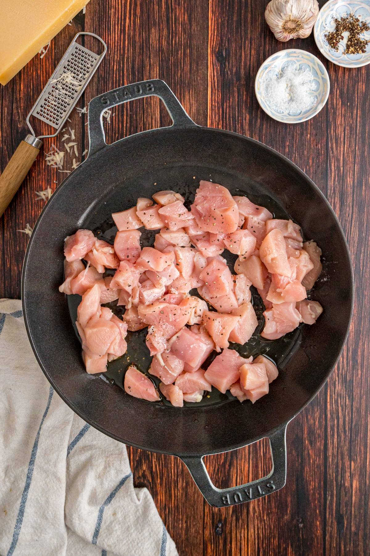 Diced raw chicken in a black skillet, surrounded by garlic, salt, pepper, and a cheese grater on a wooden table.