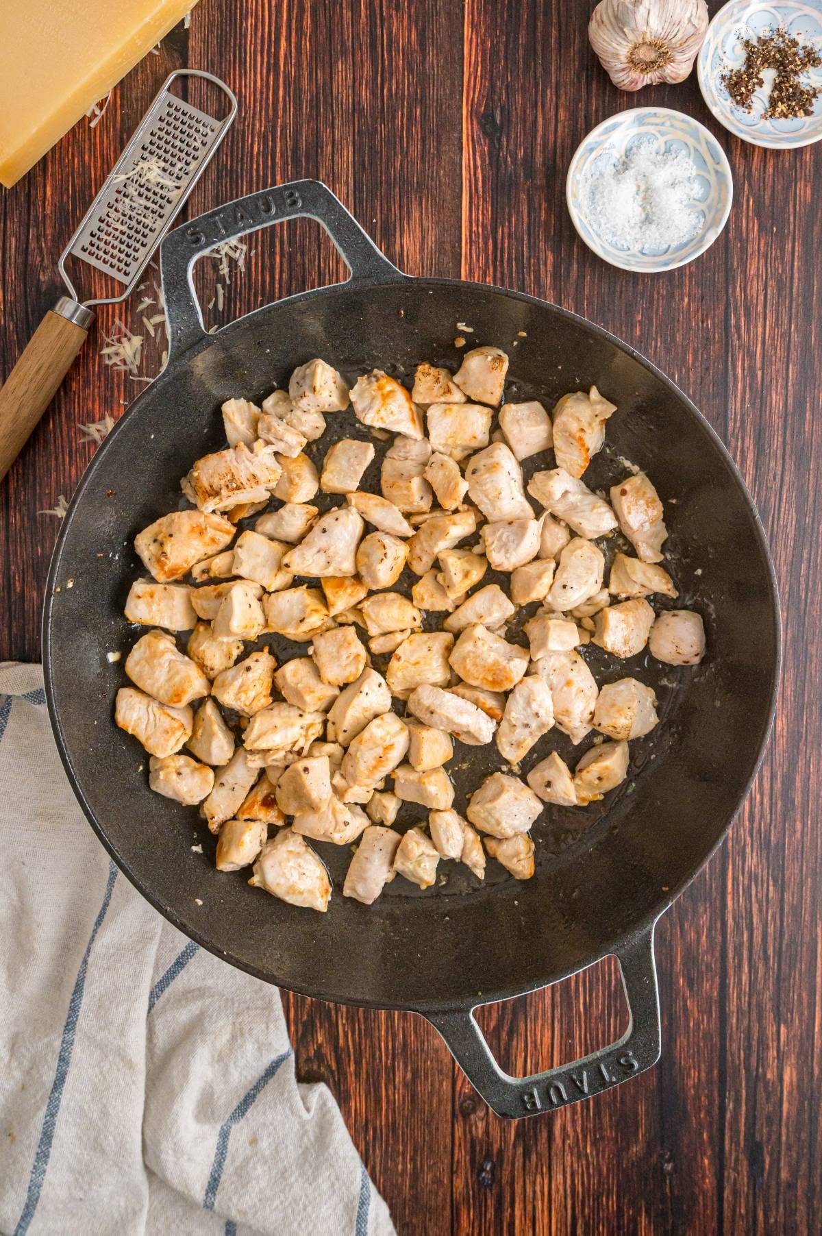 Cubed chicken cooking in a black skillet on a wooden surface, surrounded by seasonings and a cheese grater.