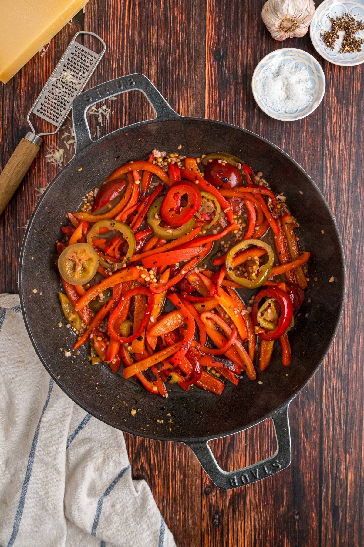 Sliced red peppers and jalapeños sautéing in a cast iron pan on a wooden table with seasonings nearby.