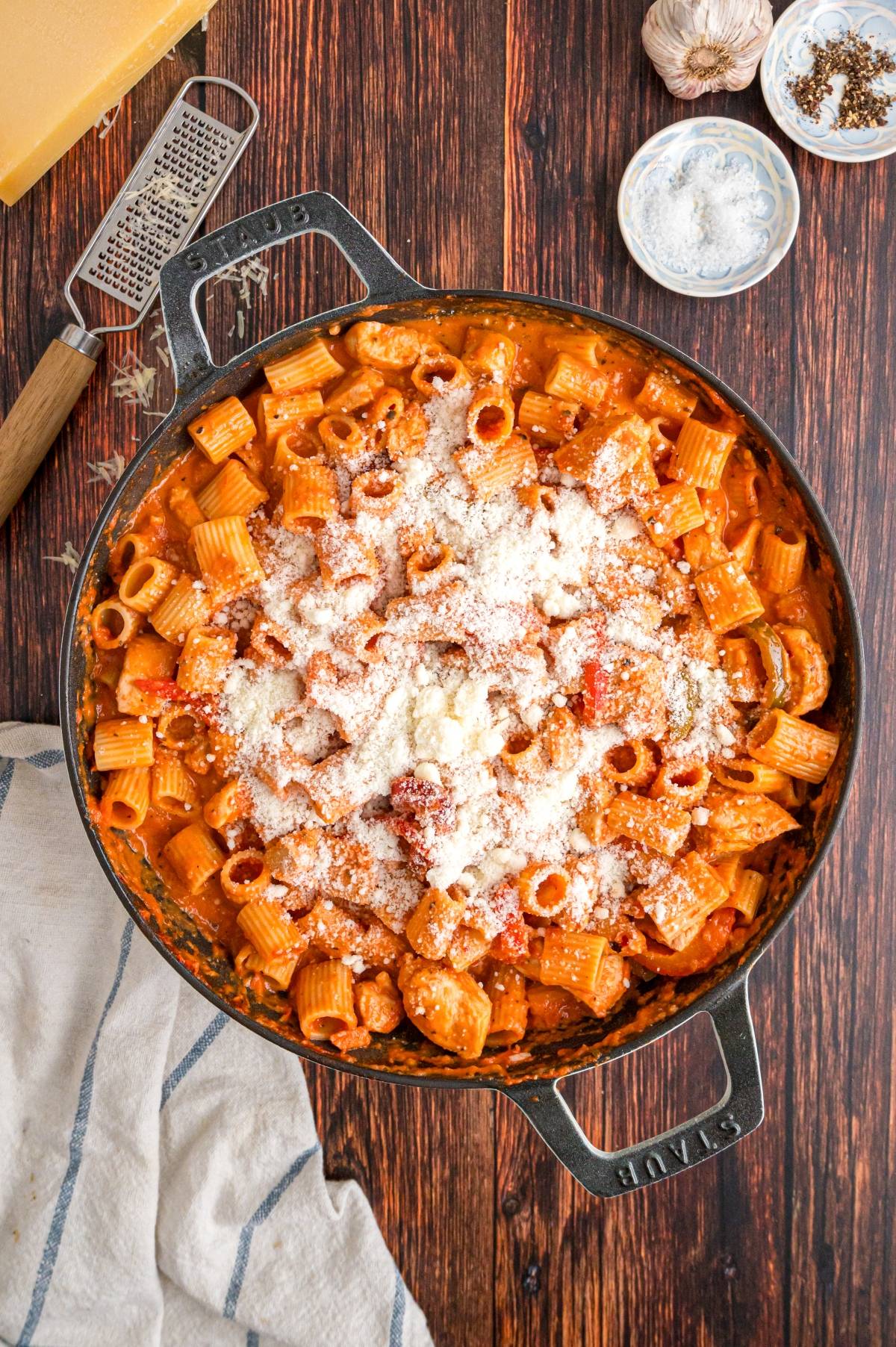 Large skillet of rigatoni pasta with red sauce and grated cheese, on a wooden table with a grater and seasonings.