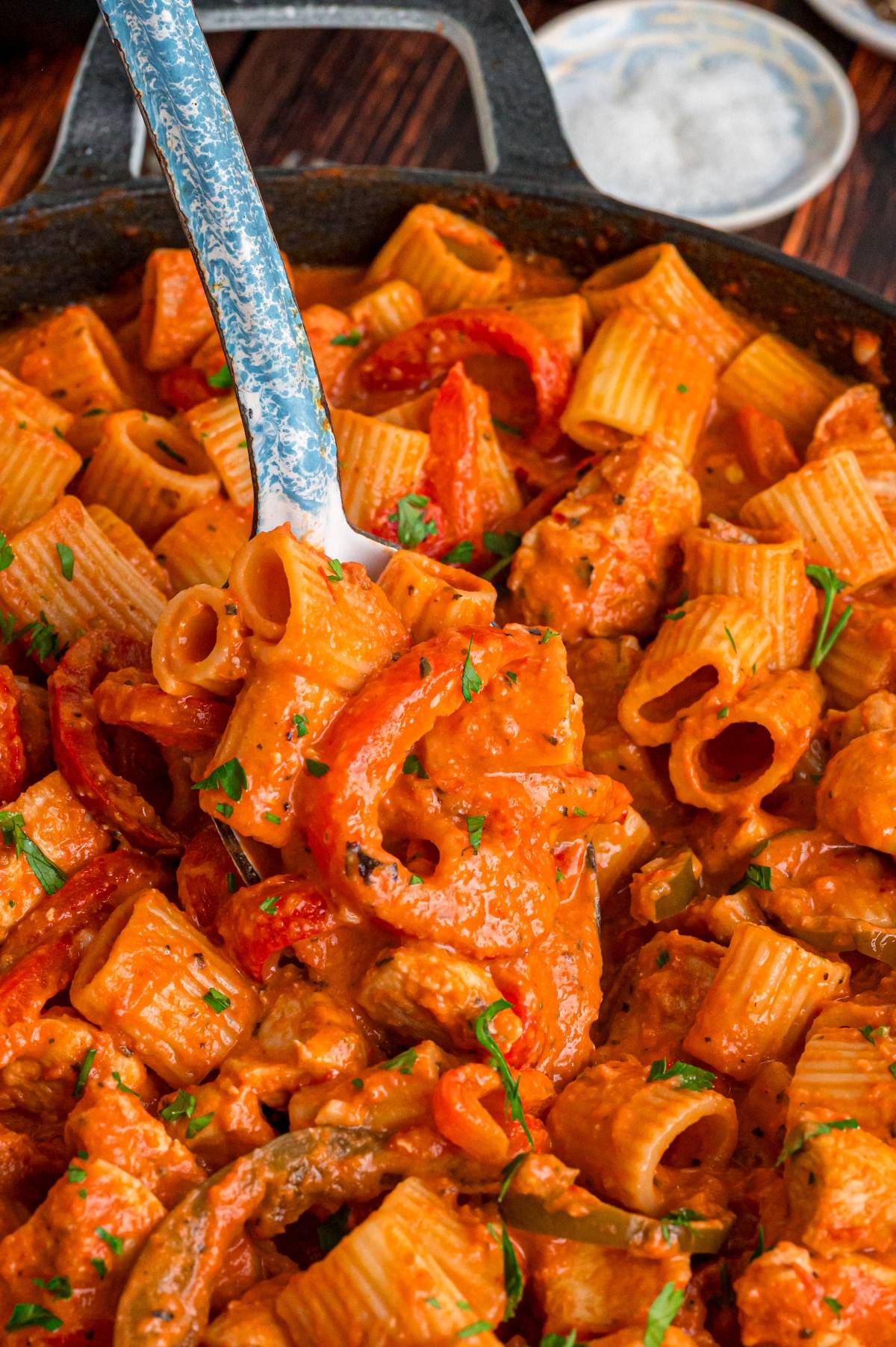 Creamy rigatoni pasta with bell peppers and herbs in a skillet, being stirred with a metal spoon.