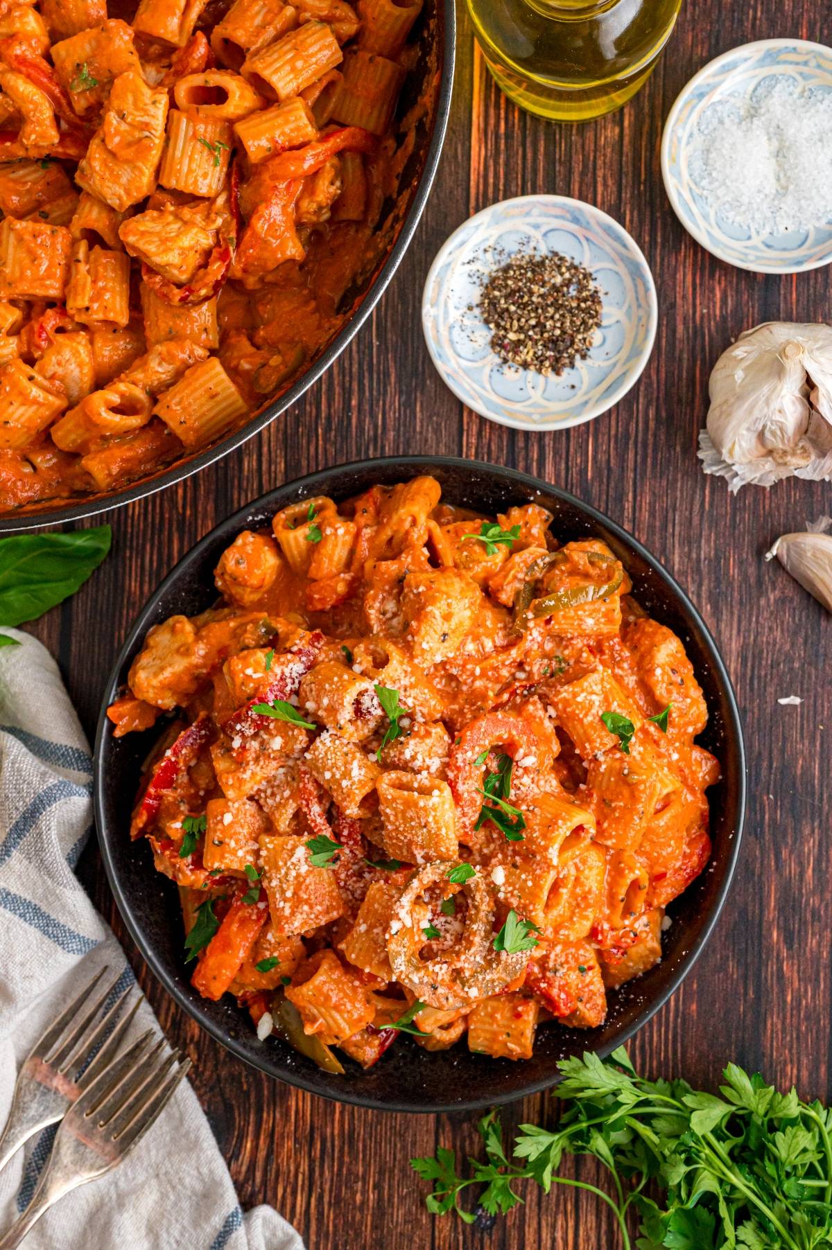 Bowl of creamy rigatoni pasta with vegetables, topped with herbs and Parmesan, next to seasoning and garlic.