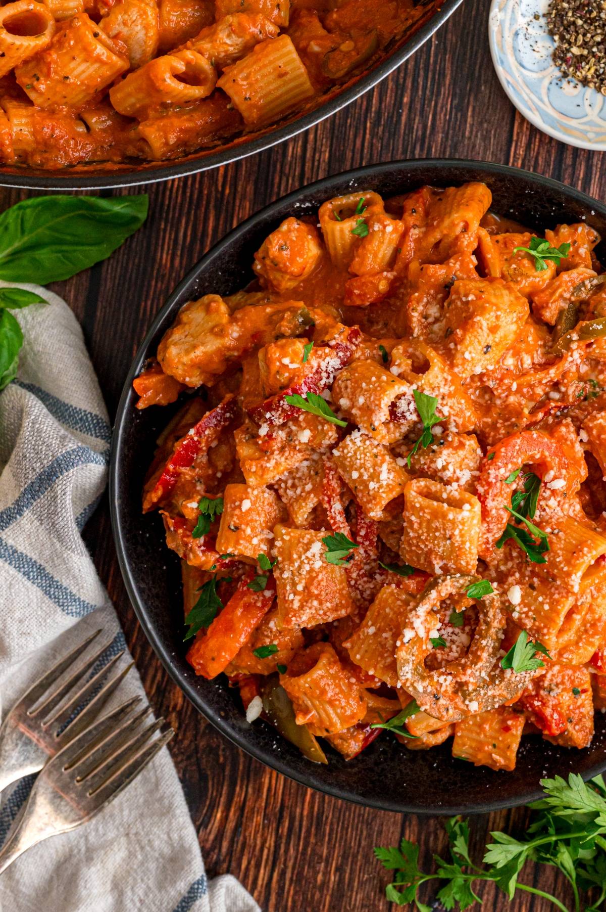 A bowl of creamy rigatoni pasta with vegetables, topped with parsley and grated cheese on a wooden table.
