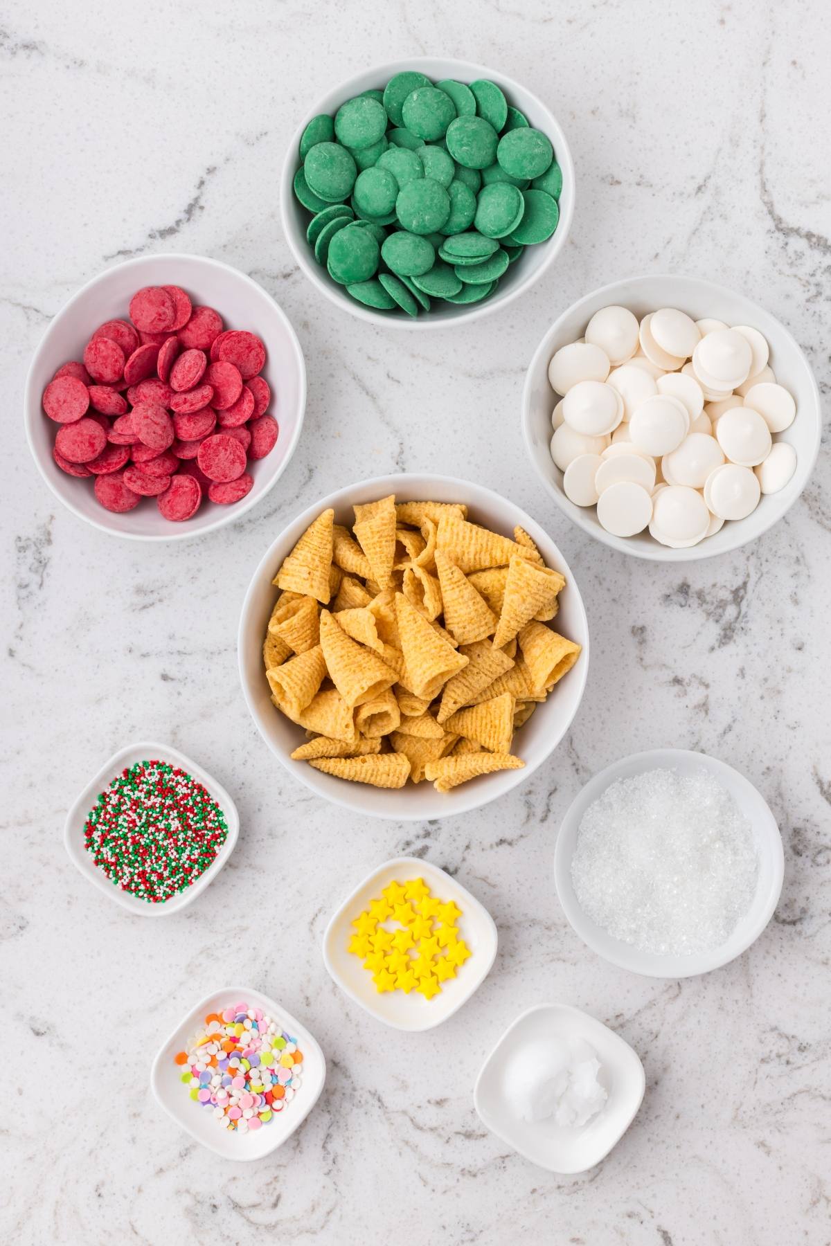 Assorted bowls of green, red, and white candy melts, Bugles, and colorful sprinkles on a marble surface.
