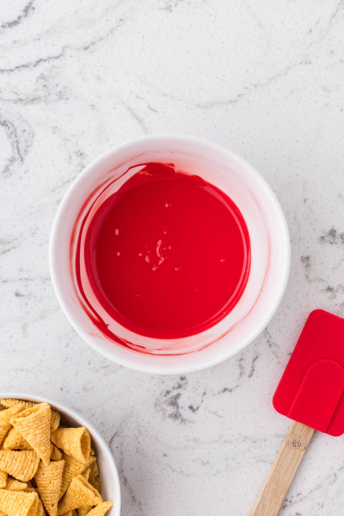 A bowl of bright red melted candy next to cereal pieces and a red spatula on a white countertop.