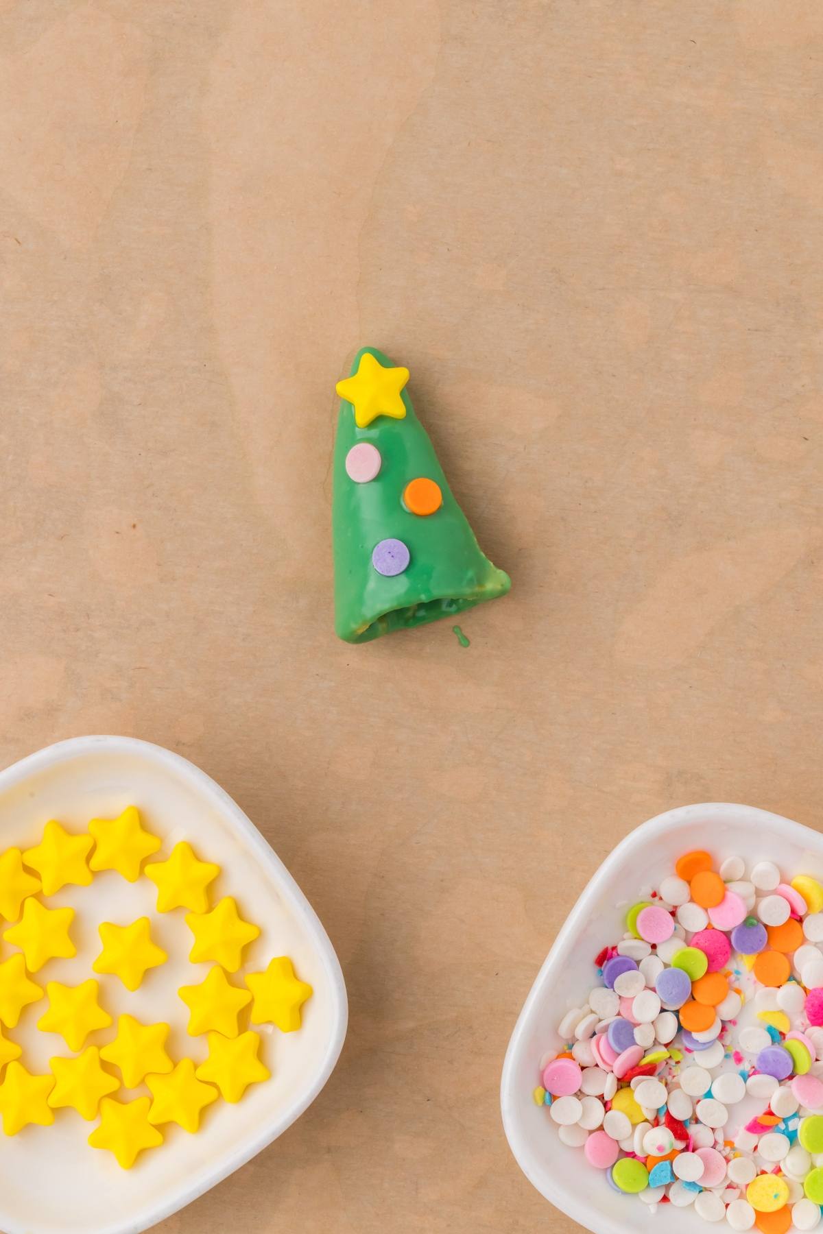 Green Christmas tree-shaped cookie with star and dot sprinkles, with bowls of star and dot sprinkles nearby.