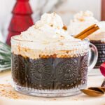 Glass mug of Christmas coffee topped with whipped cream and cinnamon, surrounded by festive holiday decorations.