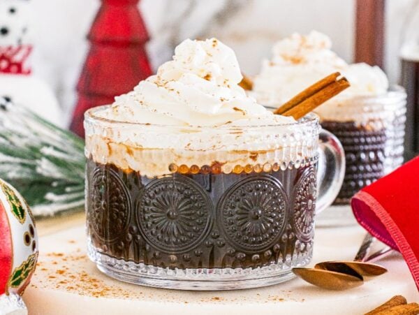 Glass mug of Christmas coffee topped with whipped cream and cinnamon, surrounded by festive holiday decorations.