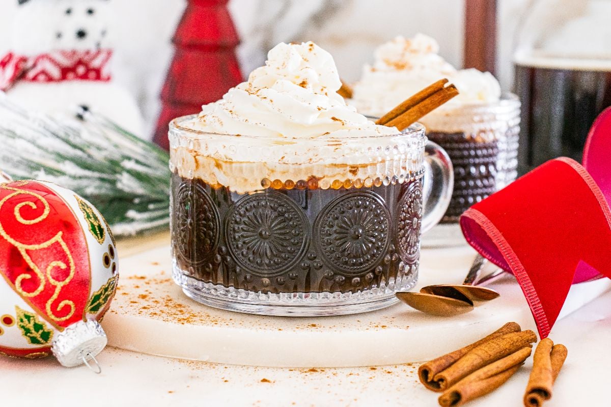 Glass mug of hot chocolate with whipped cream, a cinnamon stick, and festive holiday decorations&mdash;perfect for enjoying alongside your favorite Christmas coffee.