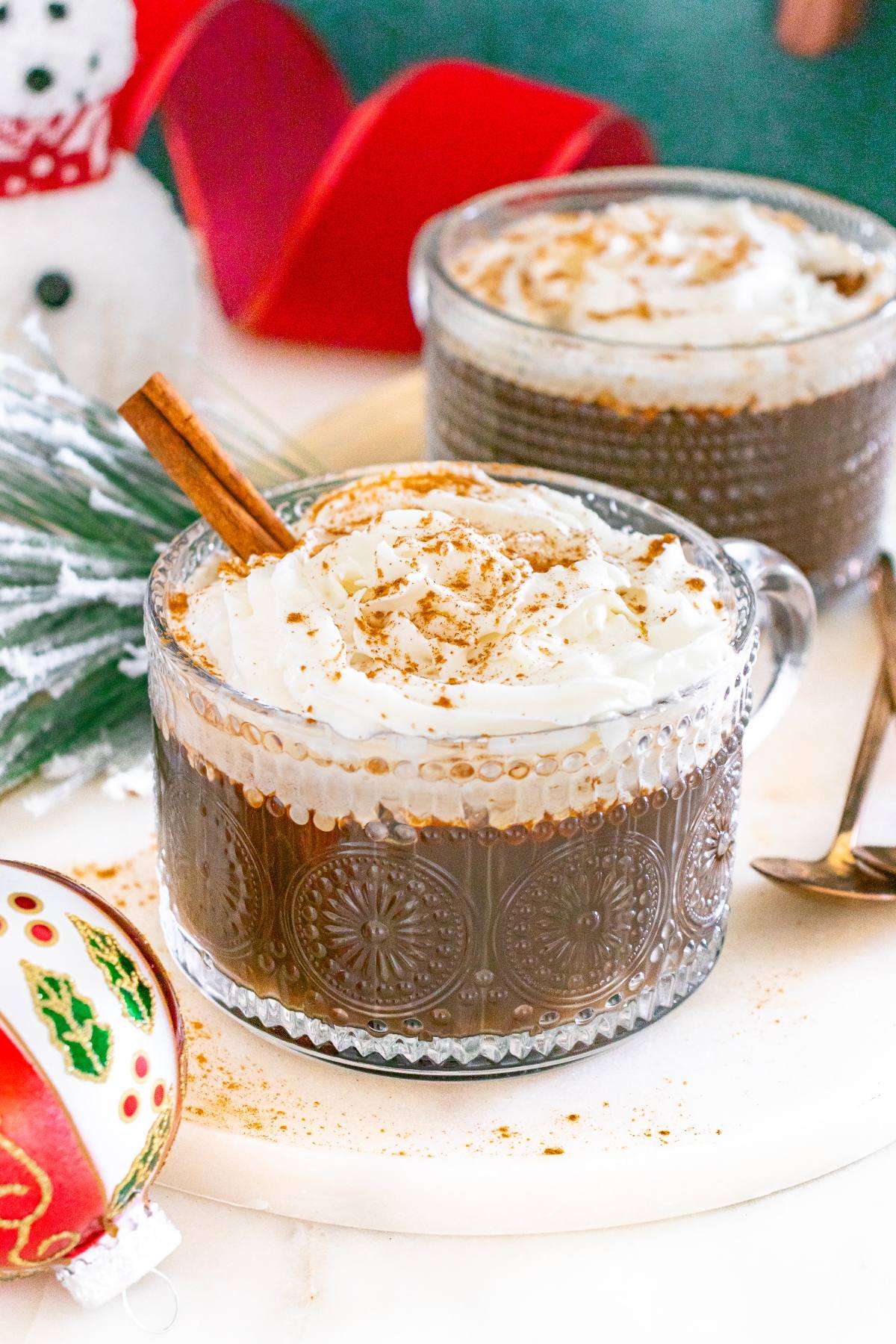 Two mugs of hot chocolate topped with whipped cream and cinnamon sticks, with holiday decorations nearby.