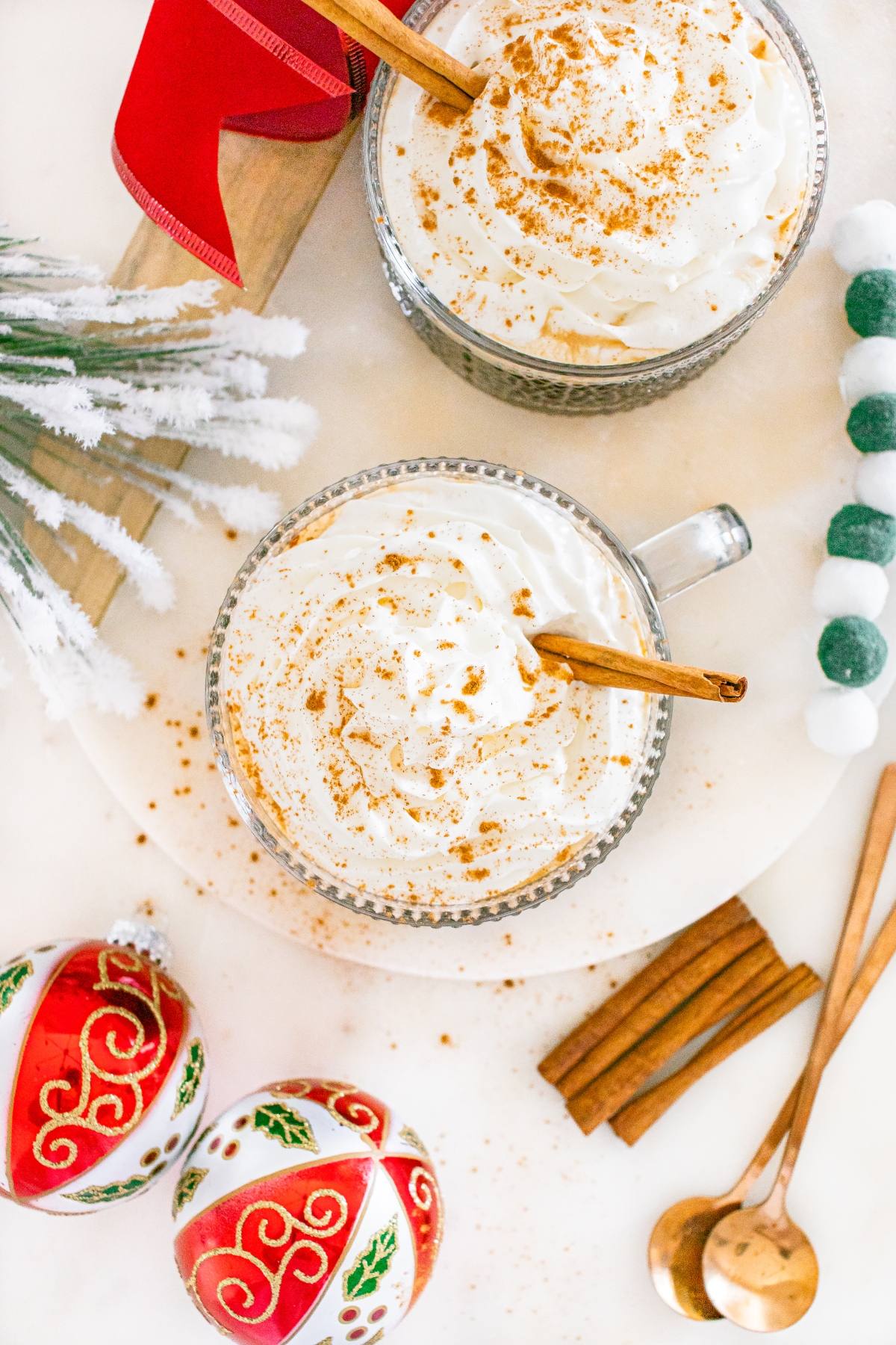 Two mugs of whipped cream-topped drinks with cinnamon, Christmas ornaments, and festive decorations nearby.