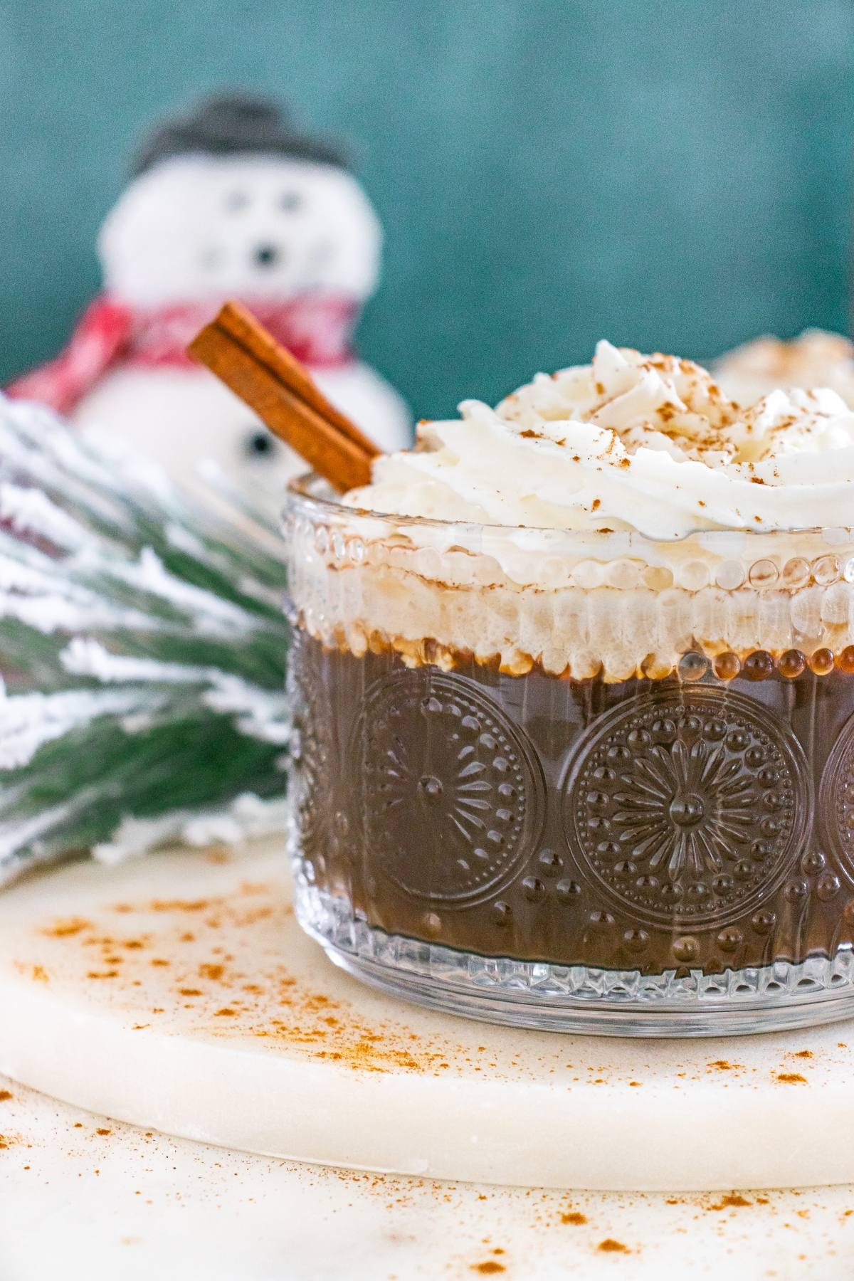 A glass of hot chocolate with whipped cream and a cinnamon stick, with a snowman decoration in the background.