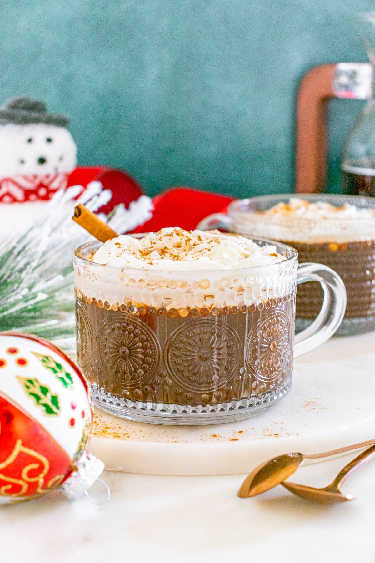 A glass mug of hot chocolate topped with whipped cream and cinnamon, with festive holiday decor in the background.