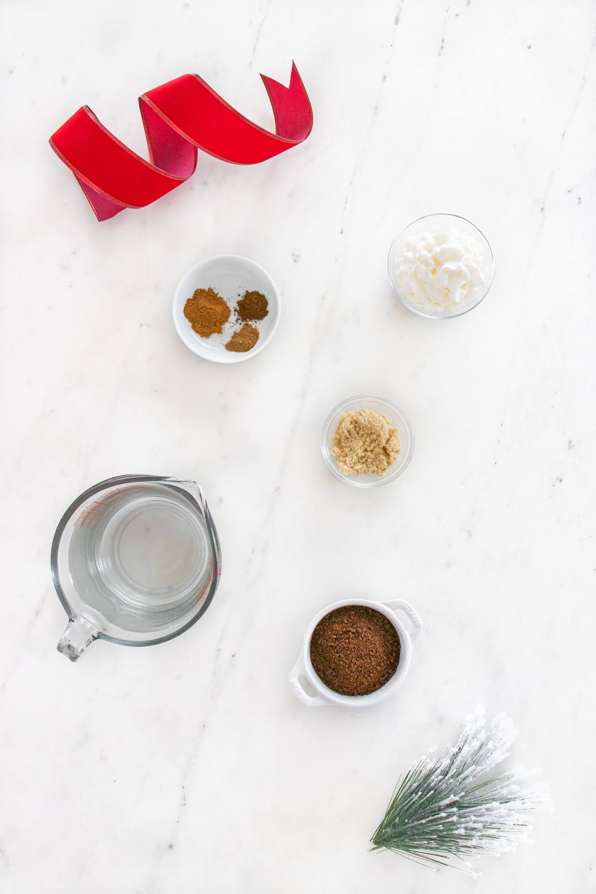 Overhead view of coffee, brown sugar, spices, whipped cream, a red ribbon, and a pine branch on a white surface.