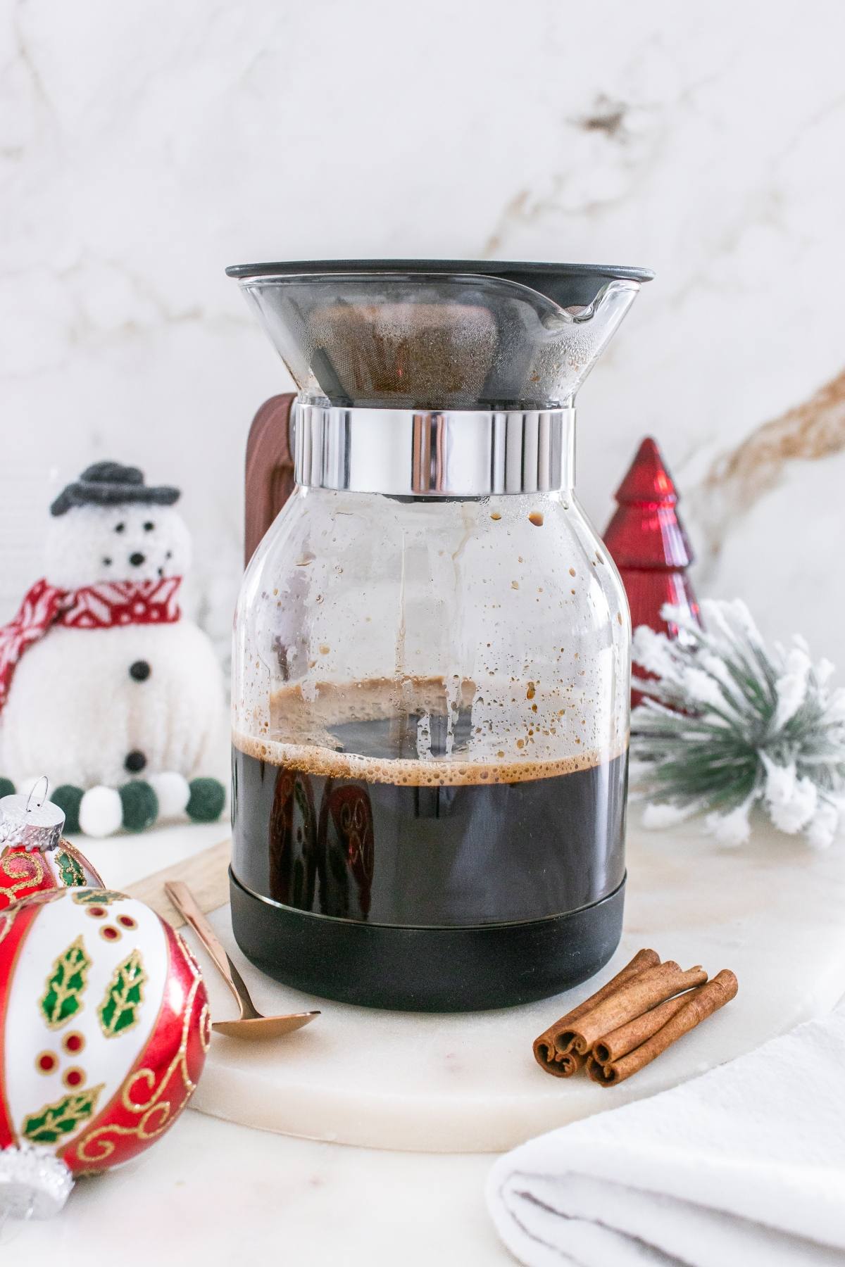 A glass coffee maker with brewed coffee sits on a marble surface, surrounded by holiday decorations and cinnamon sticks.
