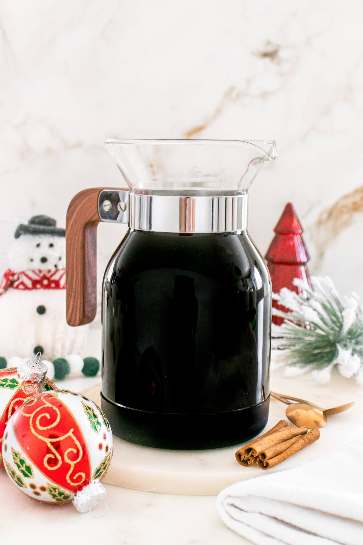 A glass carafe of black coffee with holiday decorations, a snowman, and ornaments on a white surface.
