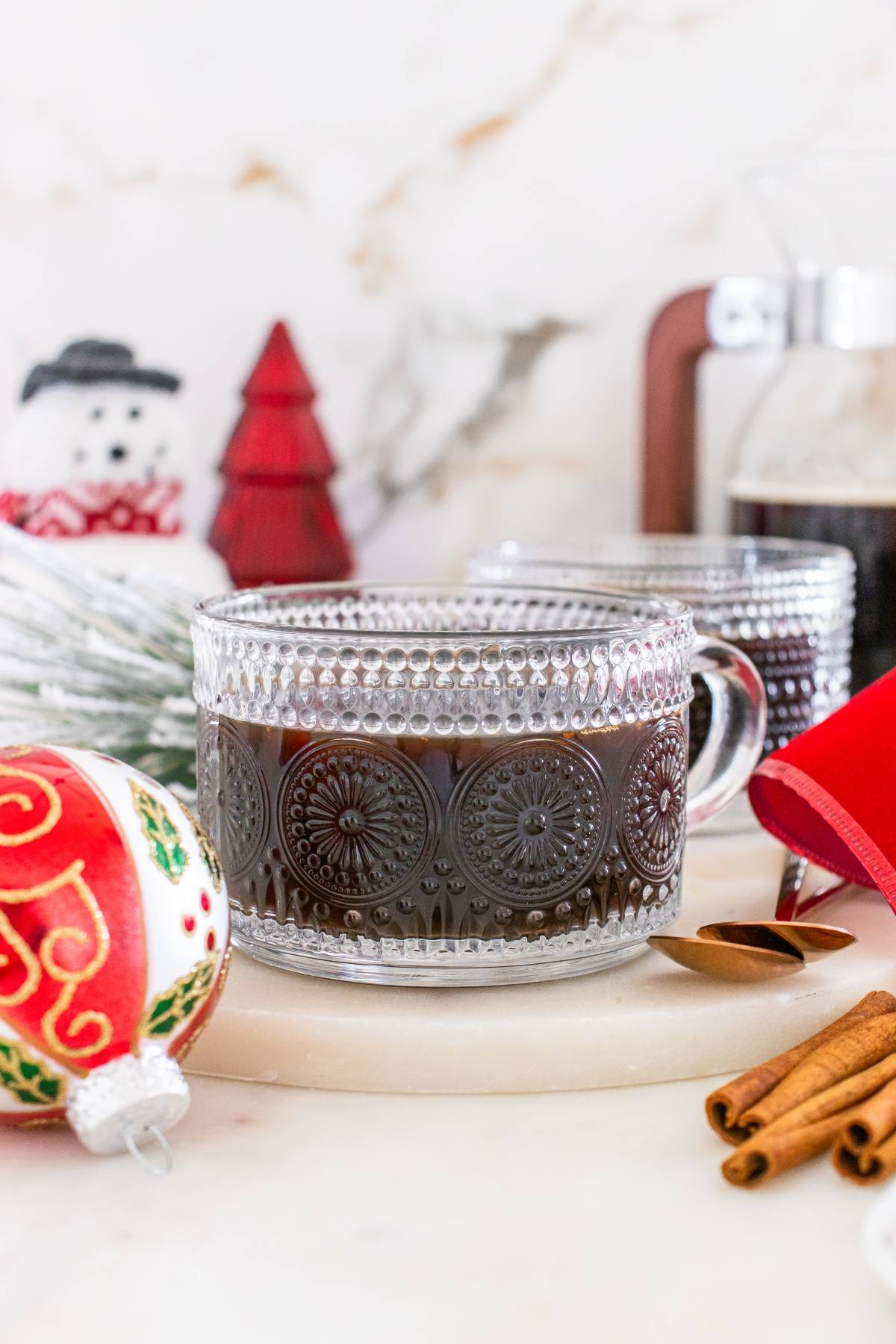 A glass mug of black coffee on a table with holiday decorations and cinnamon sticks.