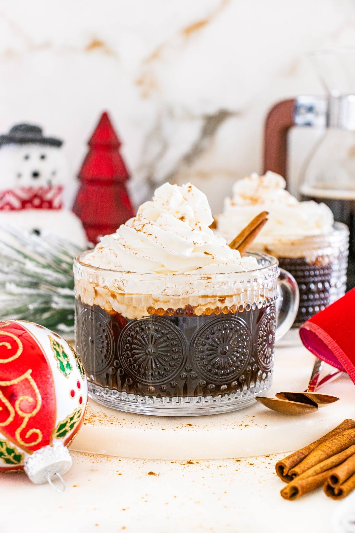 Glass mug of coffee topped with whipped cream, with Christmas ornaments and decorations in the background.
