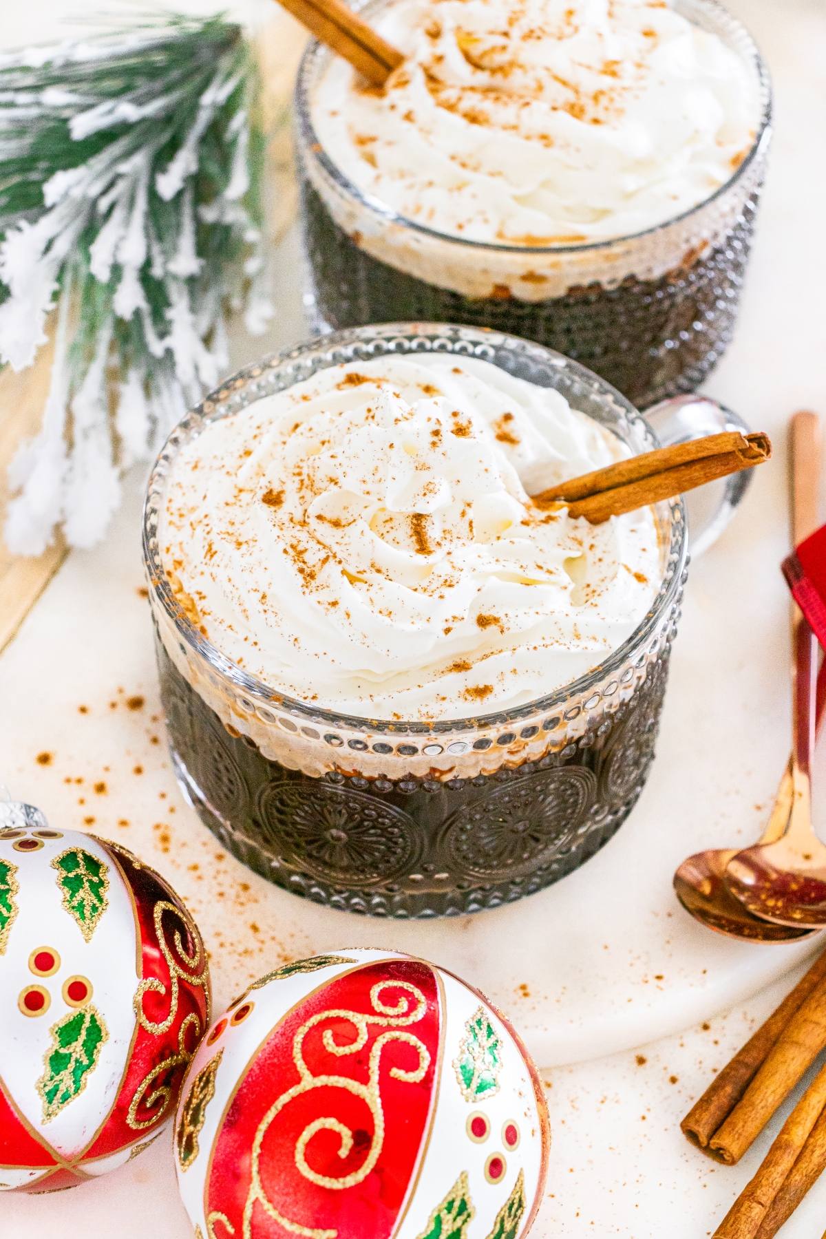 Two glass mugs of whipped cream-topped coffee with cinnamon sticks, next to holiday ornaments and festive decor.