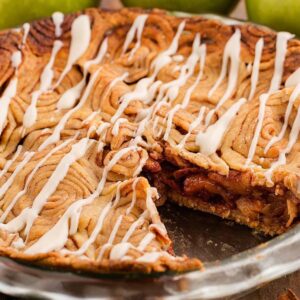 A sliced cinnamon roll apple pie with icing drizzle in a glass dish, surrounded by green apples on a wooden table.