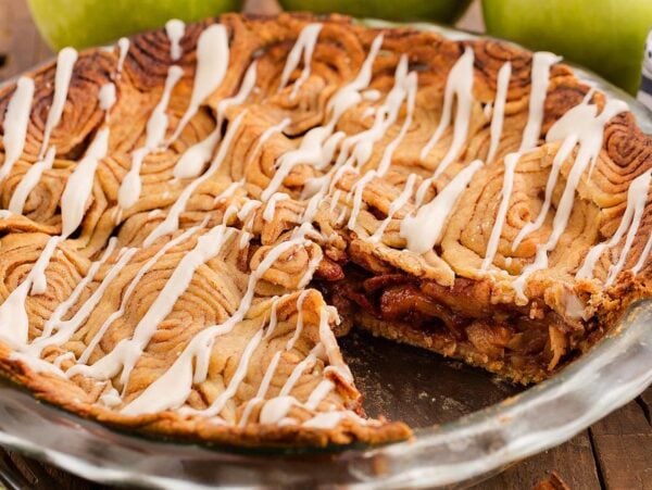 A sliced cinnamon roll apple pie with icing drizzle in a glass dish, surrounded by green apples on a wooden table.