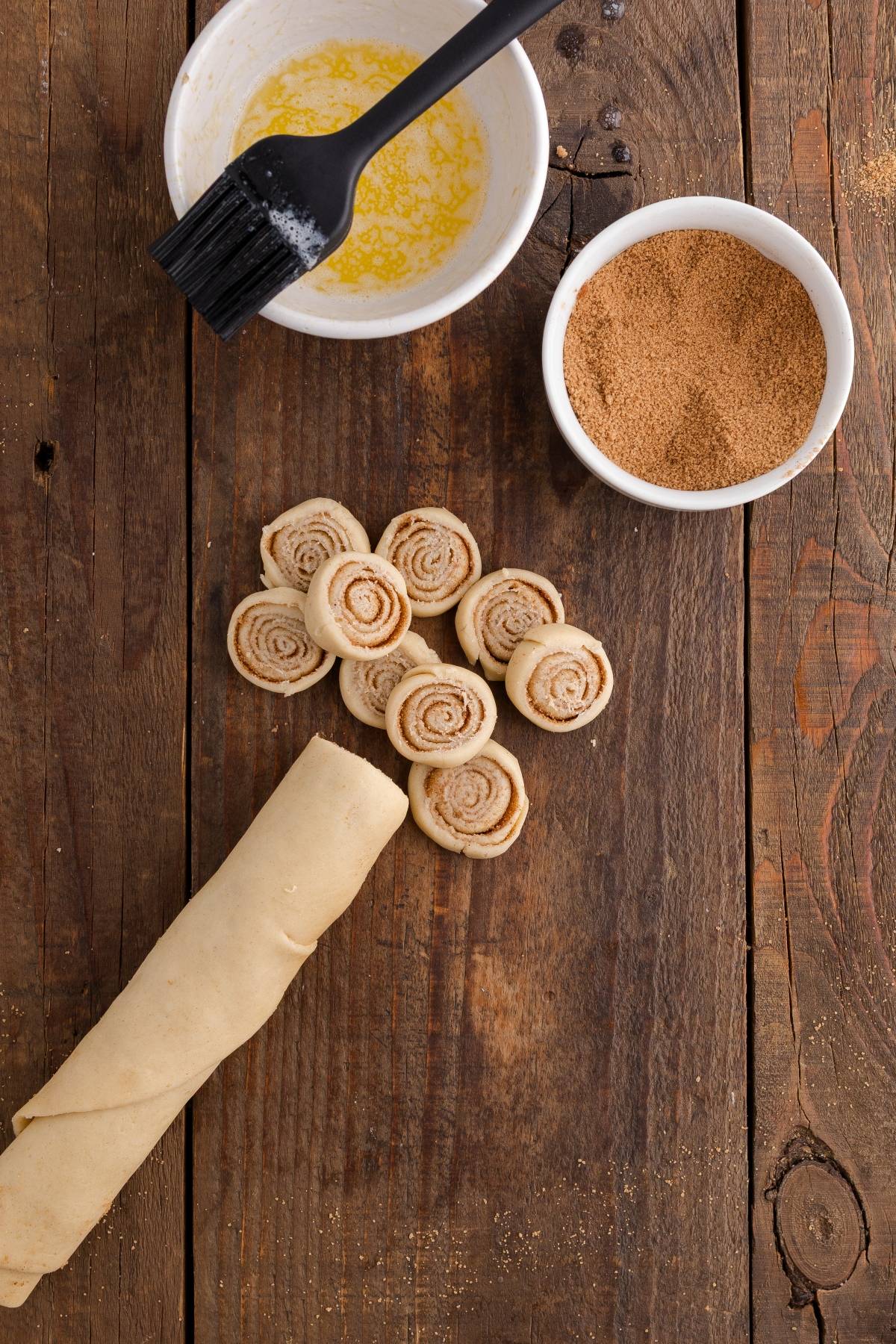 Sliced cinnamon roll dough, melted butter, and cinnamon sugar on a rustic wooden table.