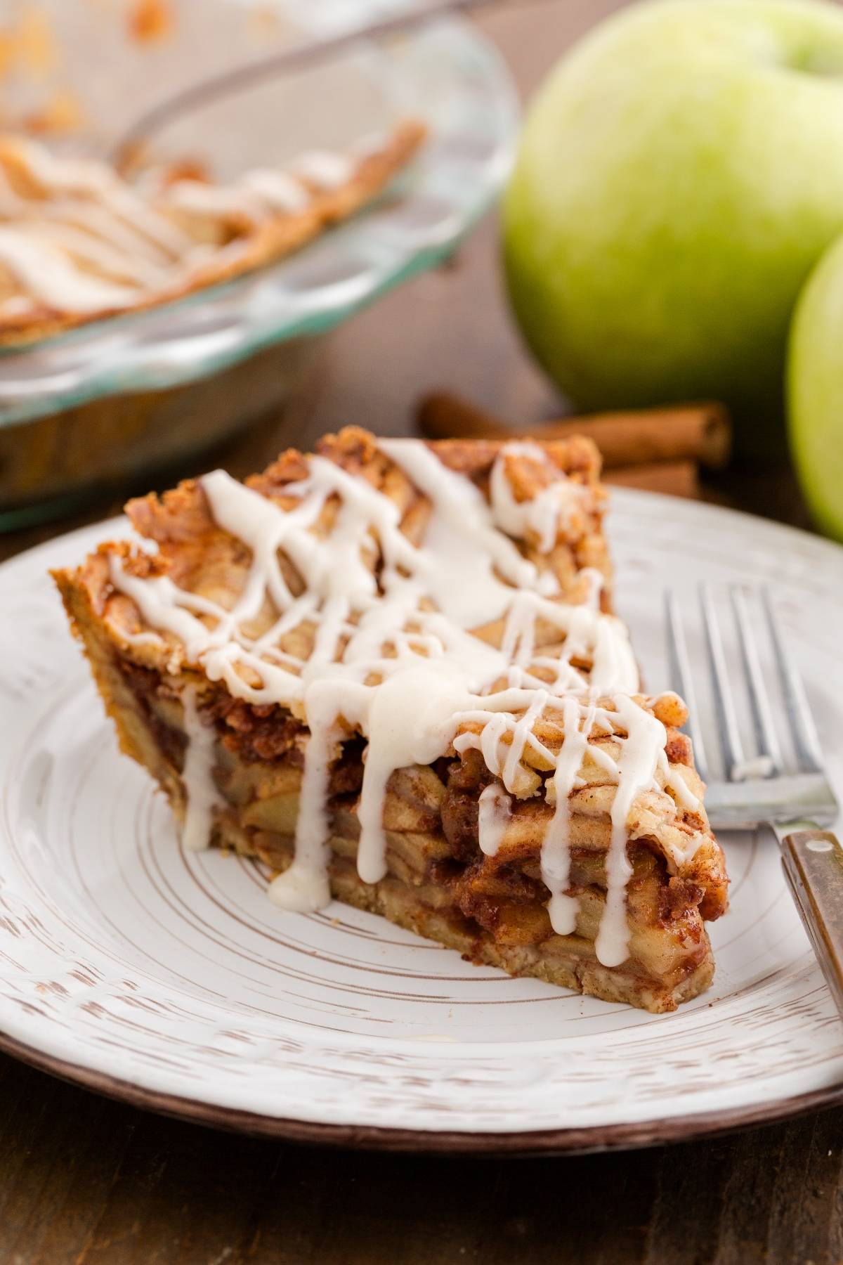 A slice of apple pie with white icing on a plate, next to green apples and a fork.
