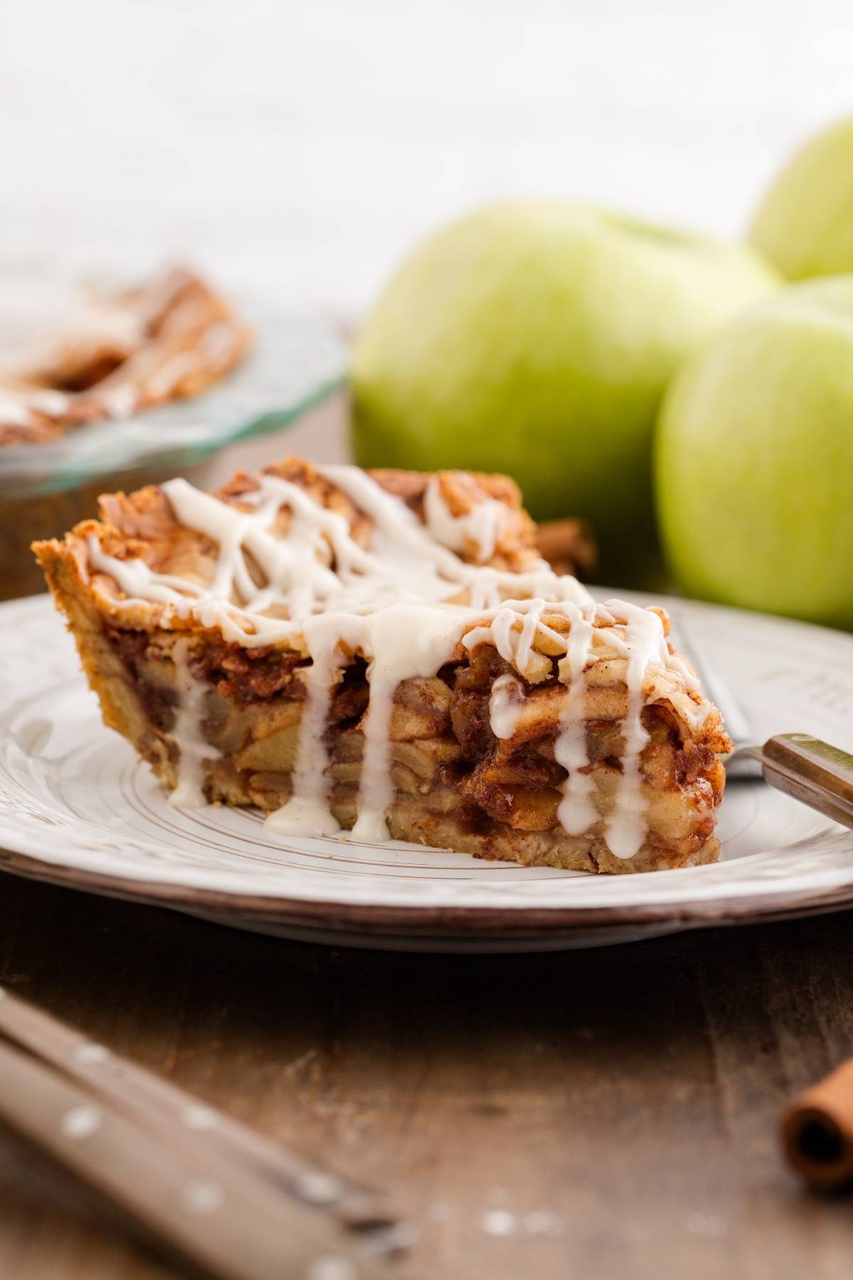 A slice of apple pie with icing drizzle on a plate, with green apples in the background.