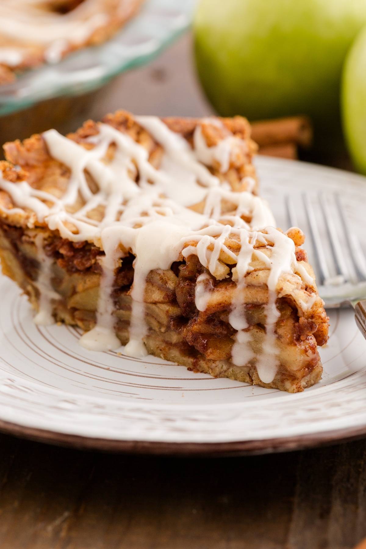 A slice of apple pie with icing drizzled on top, served on a white plate with apples in the background.