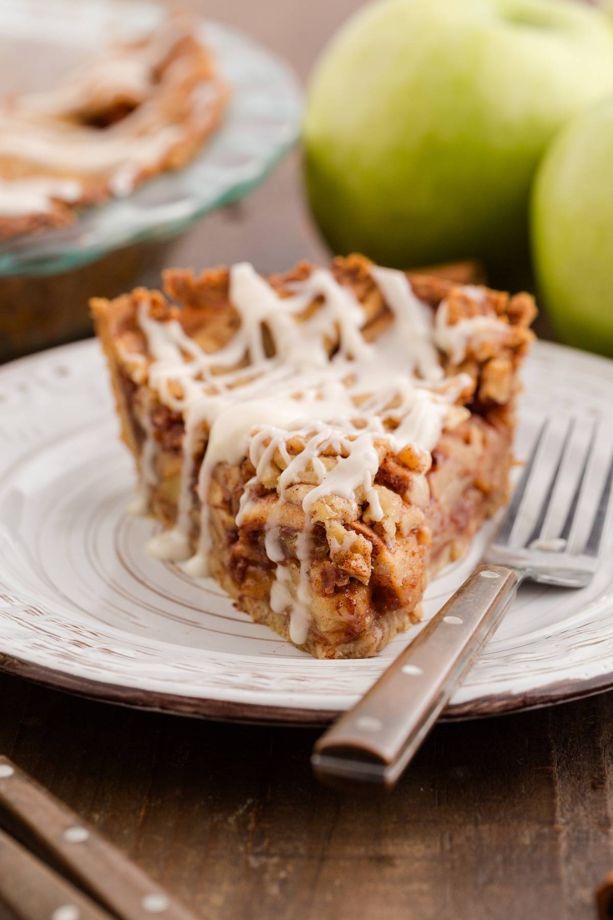 A slice of apple pie with icing drizzle on a plate, with apples and a fork beside it.