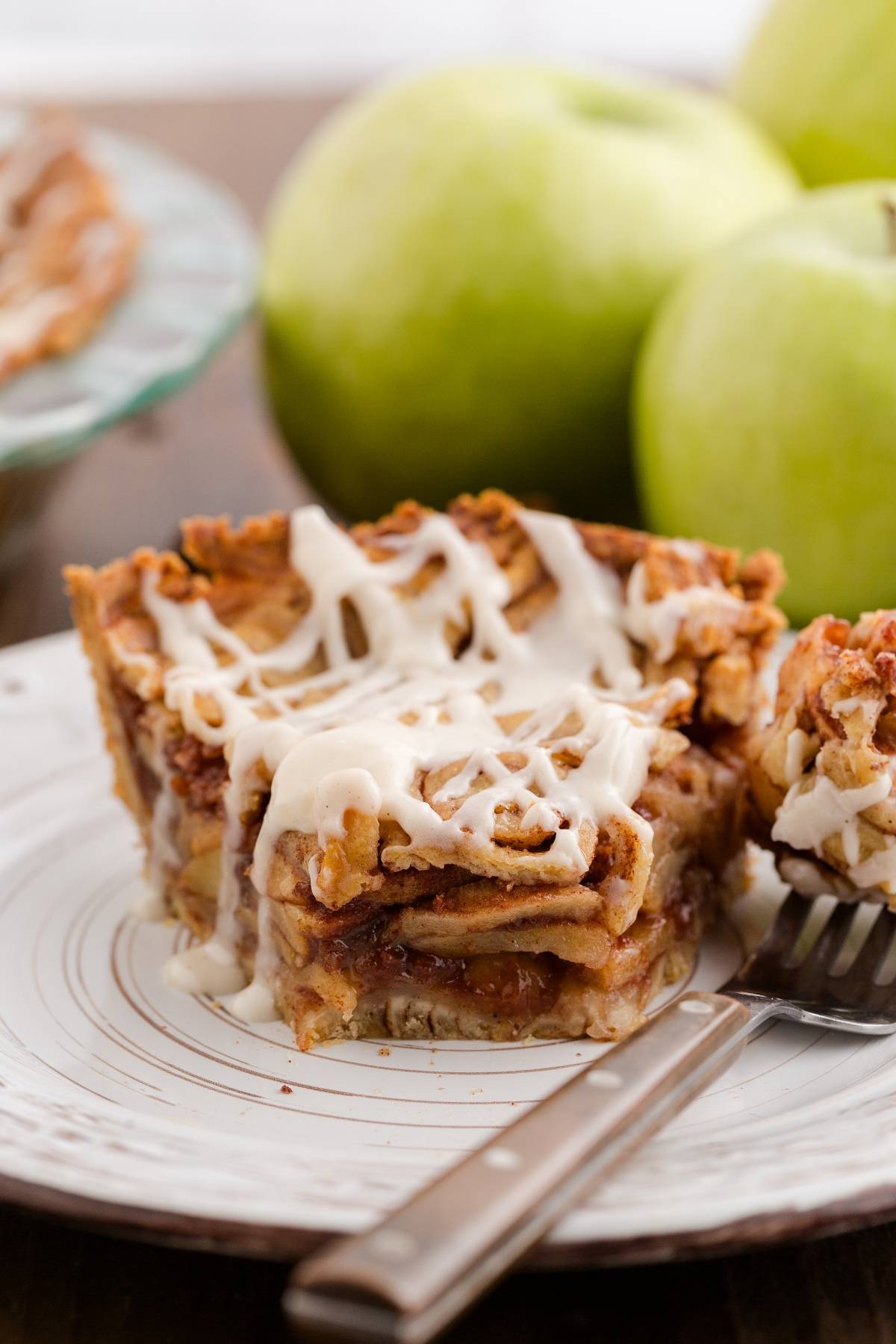 A slice of apple pie with icing on a plate, with green apples in the background and a fork beside it.