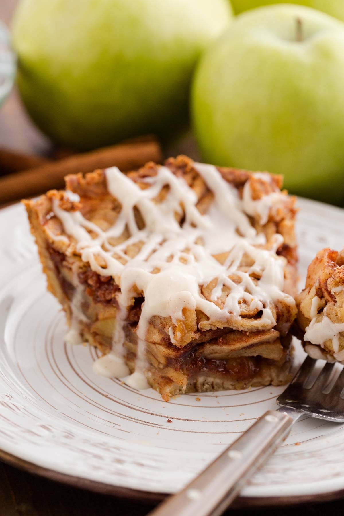 A slice of apple pie with icing on a plate, next to green apples and a fork.